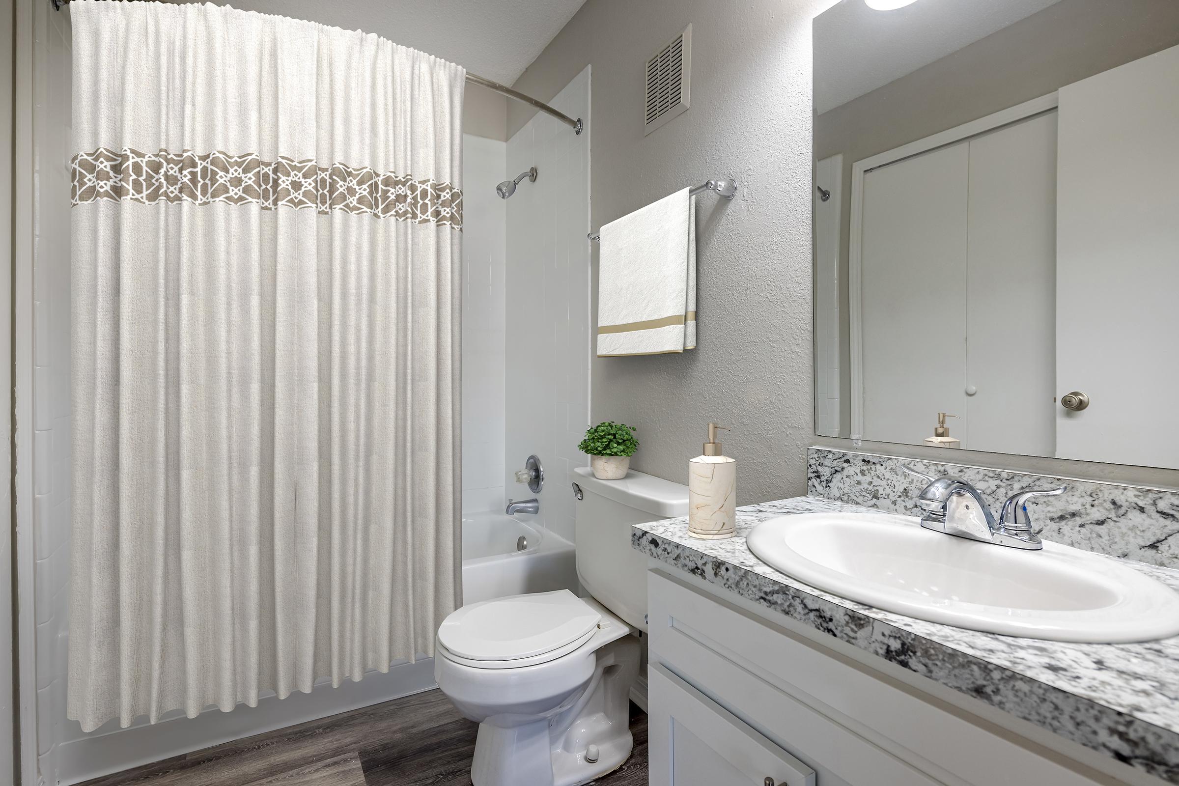 A modern bathroom featuring a white shower curtain with a decorative band, a toilet, and a sink with a granite countertop. A small potted plant is on the counter, and there is a hand towel hanging nearby. The walls are a neutral color, creating a clean and inviting atmosphere.