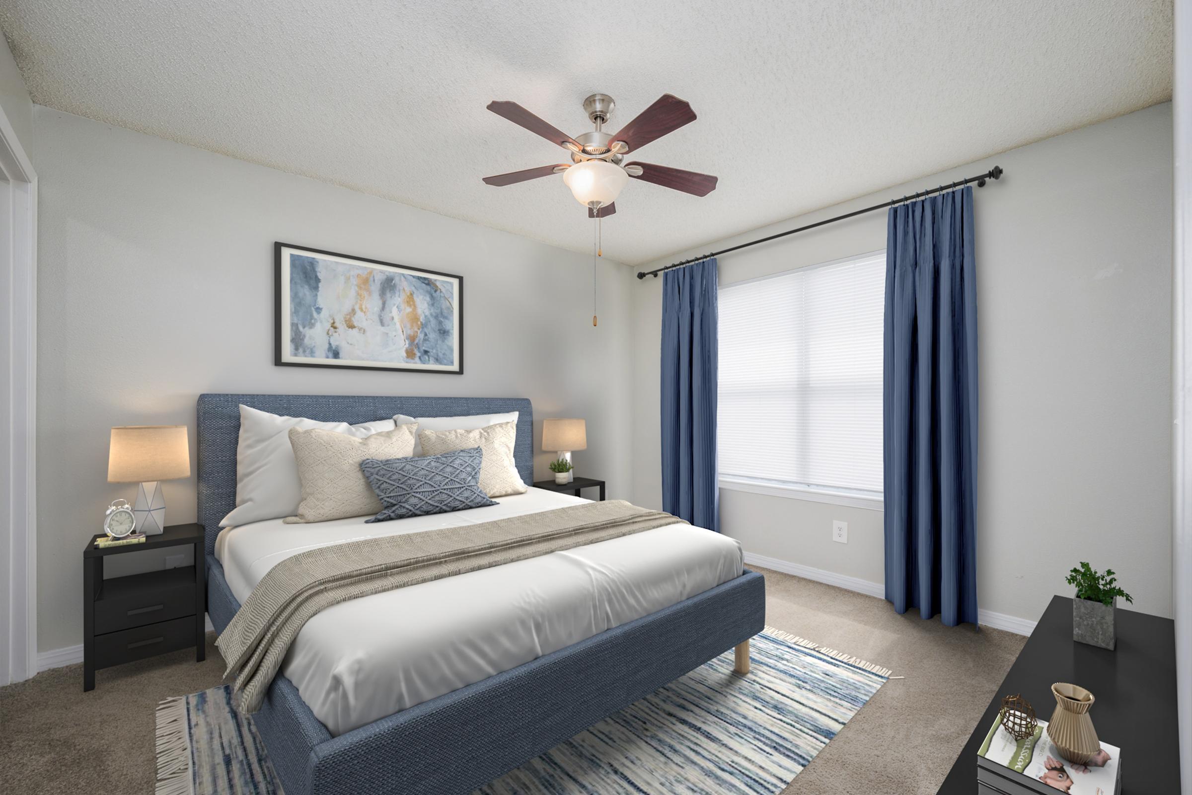A cozy bedroom featuring a queen-sized bed with white bedding and blue accents, flanked by two bedside lamps. A ceiling fan is above, and a large window with sheer curtains allows natural light. A piece of abstract art hangs on the wall, and a dark nightstand and dresser complete the decor. Soft beige carpet covers the floor.