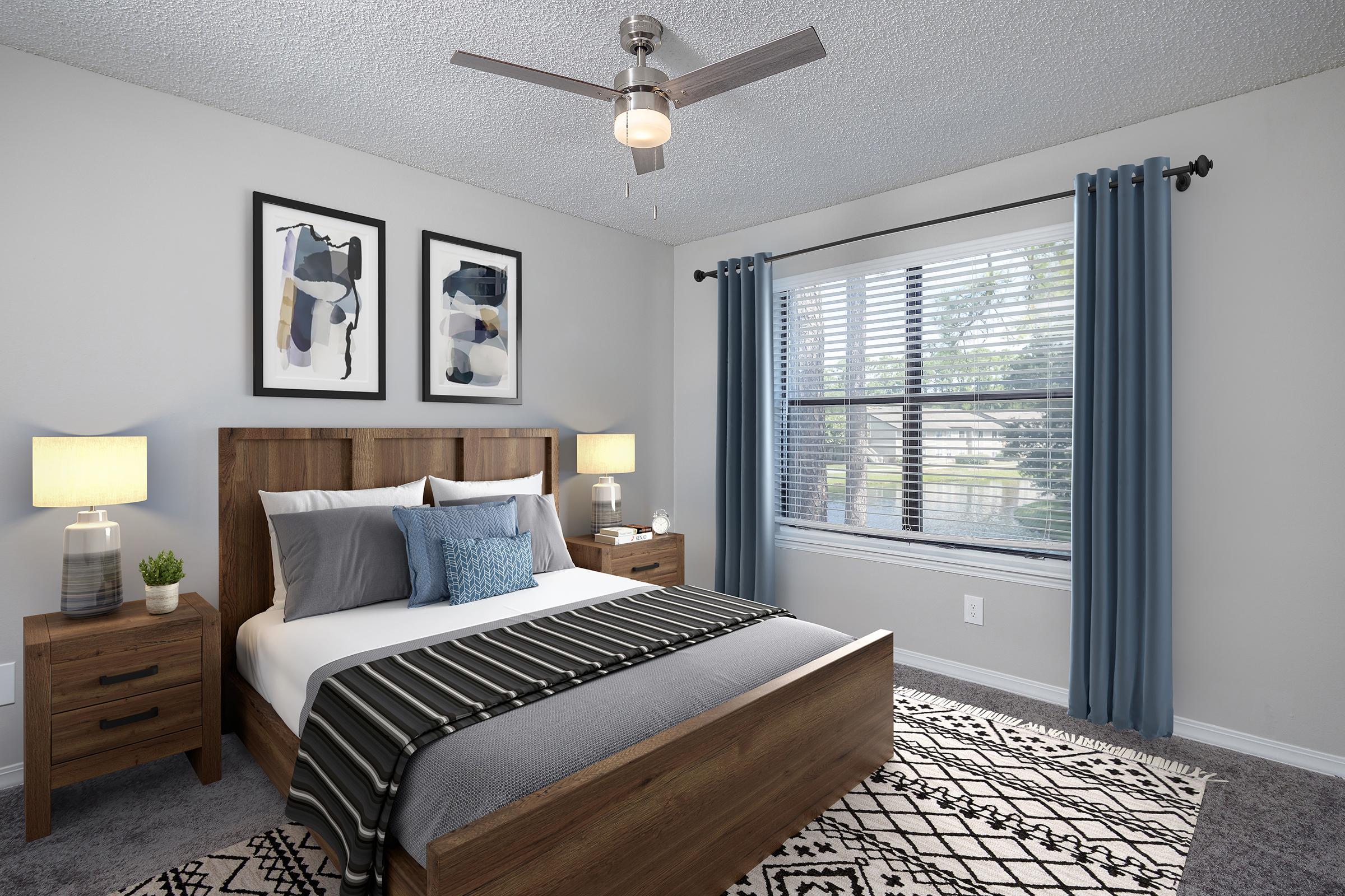 A modern bedroom featuring a wooden bed frame with gray bedding and decorative pillows. Two nightstands with lamps are on either side, and the walls are adorned with abstract art. A ceiling fan is visible, and large windows with blue curtains allow natural light to brighten the space, showcasing a view of the outdoors.