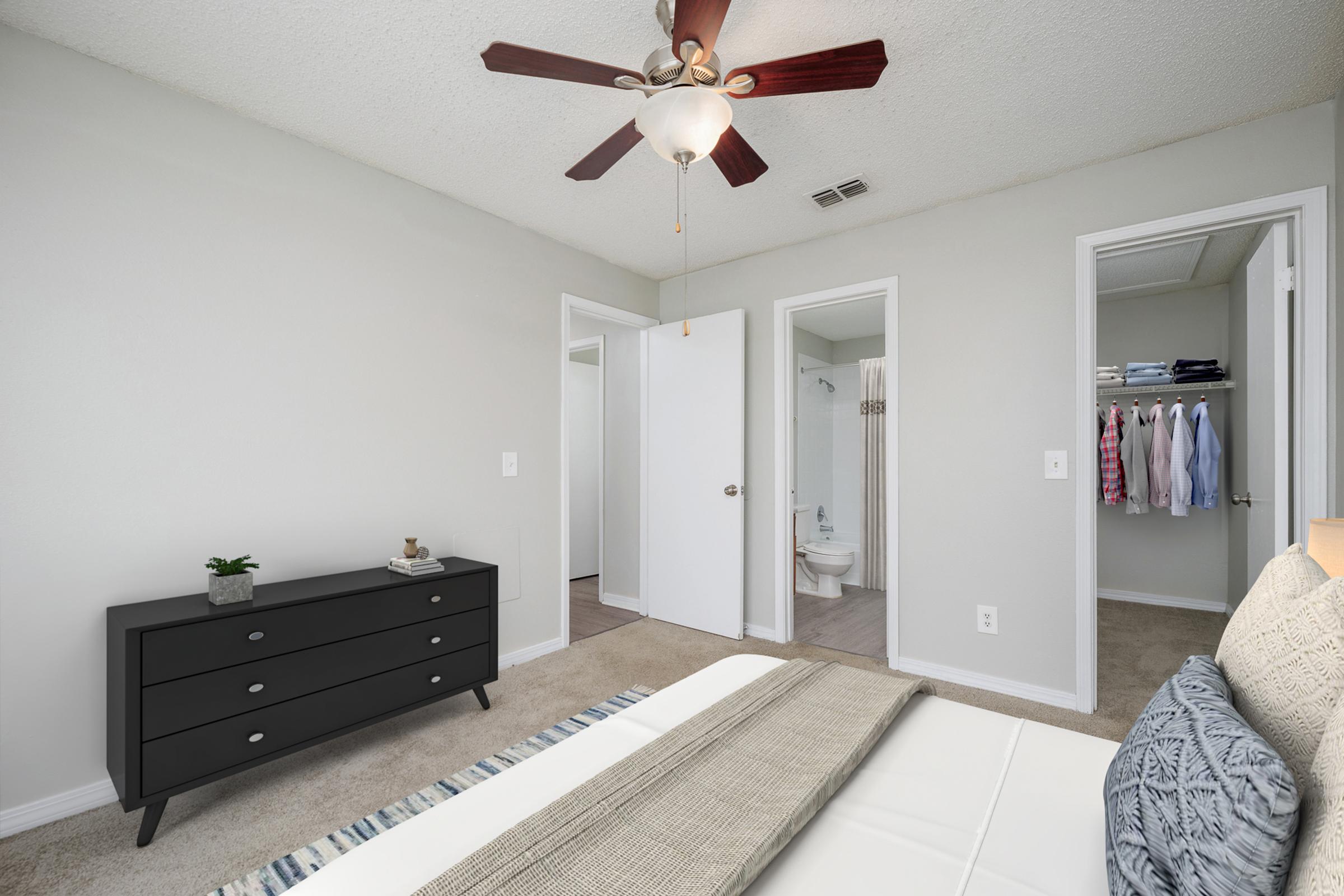 A well-lit bedroom featuring a modern dresser with drawers, a neutral color scheme, and a ceiling fan. Two open doors lead to a bathroom and another room, while a bed adorned with a cozy blanket is positioned in the foreground. The carpeted floor adds warmth to the inviting space.