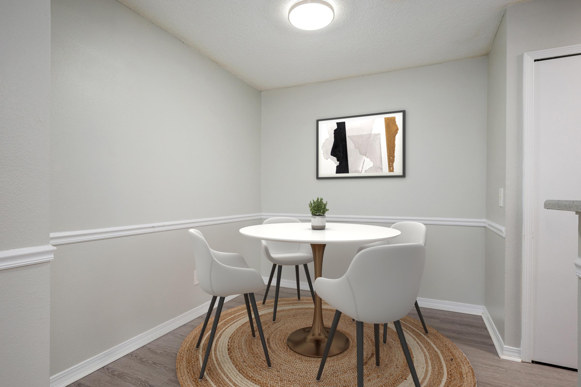 A modern dining area featuring a round white table with four white chairs on a textured circular rug. A small potted plant sits on the table. The light-gray walls are accented by white wainscoting, and a framed abstract artwork hangs on the wall above the table. A ceiling light brightens the space.