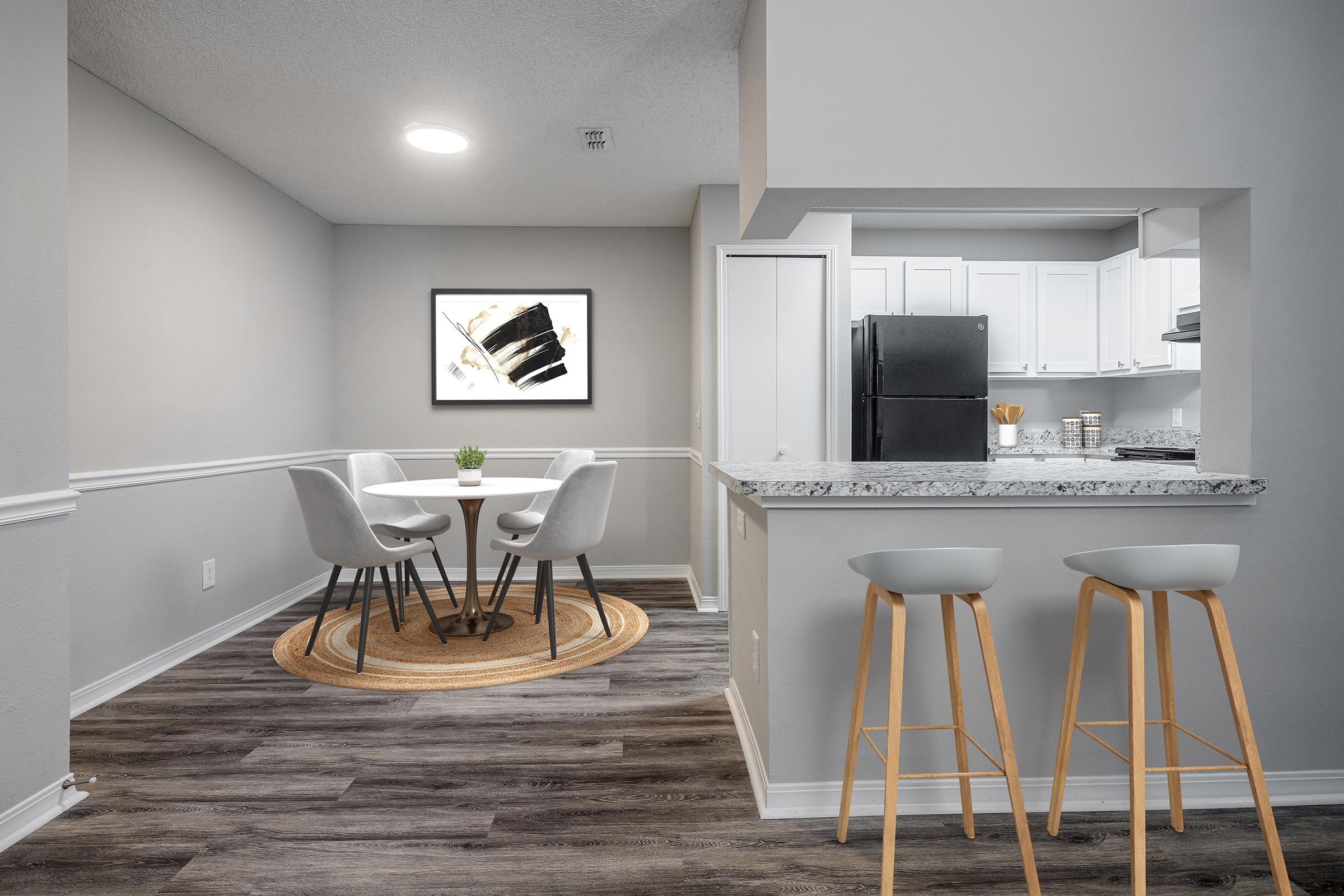 A modern kitchen and dining area with a round table surrounded by four white chairs. In the background, there is a kitchen bar with stools, a black refrigerator, and white cabinetry. A piece of abstract art is displayed on the wall, and the floor has a natural wood appearance, enhancing the contemporary style of the space.