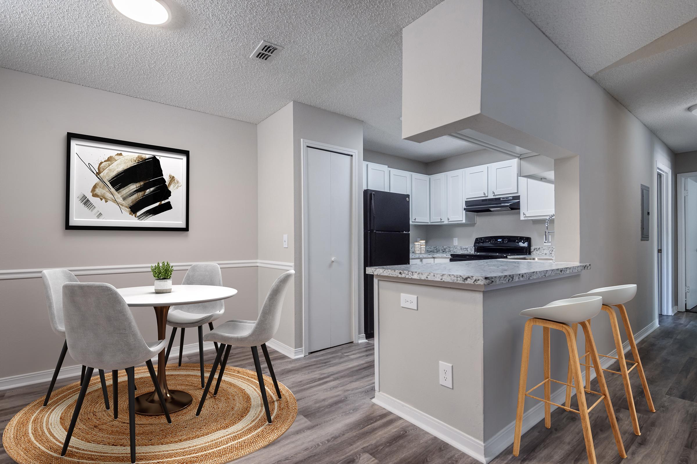 A modern kitchen and dining area featuring a round table with four grey chairs, a light-colored rug, and a framed abstract artwork on the wall. The kitchen showcases white cabinetry, a black refrigerator, and a bar with two wooden stools. The space is well-lit with a neutral color scheme.