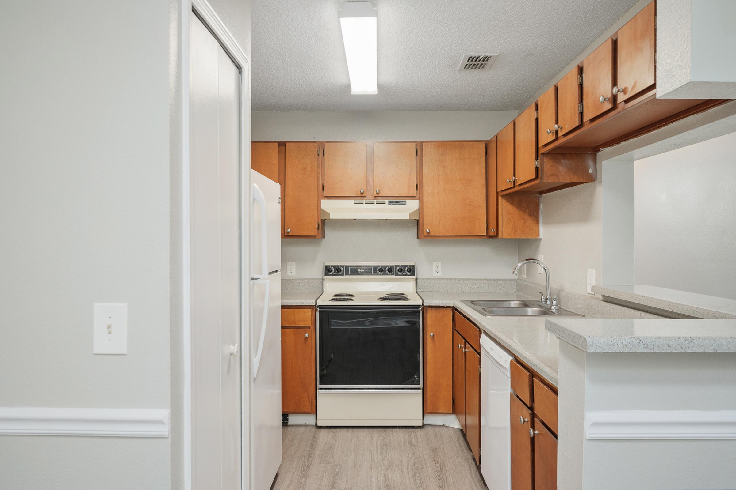 A modern kitchen featuring wooden cabinets, white countertops, an oven, and a refrigerator. The space is well-lit with overhead lighting and includes a sink and a dishwasher, showcasing a functional layout. The walls are painted in a light color, enhancing the bright and airy atmosphere.