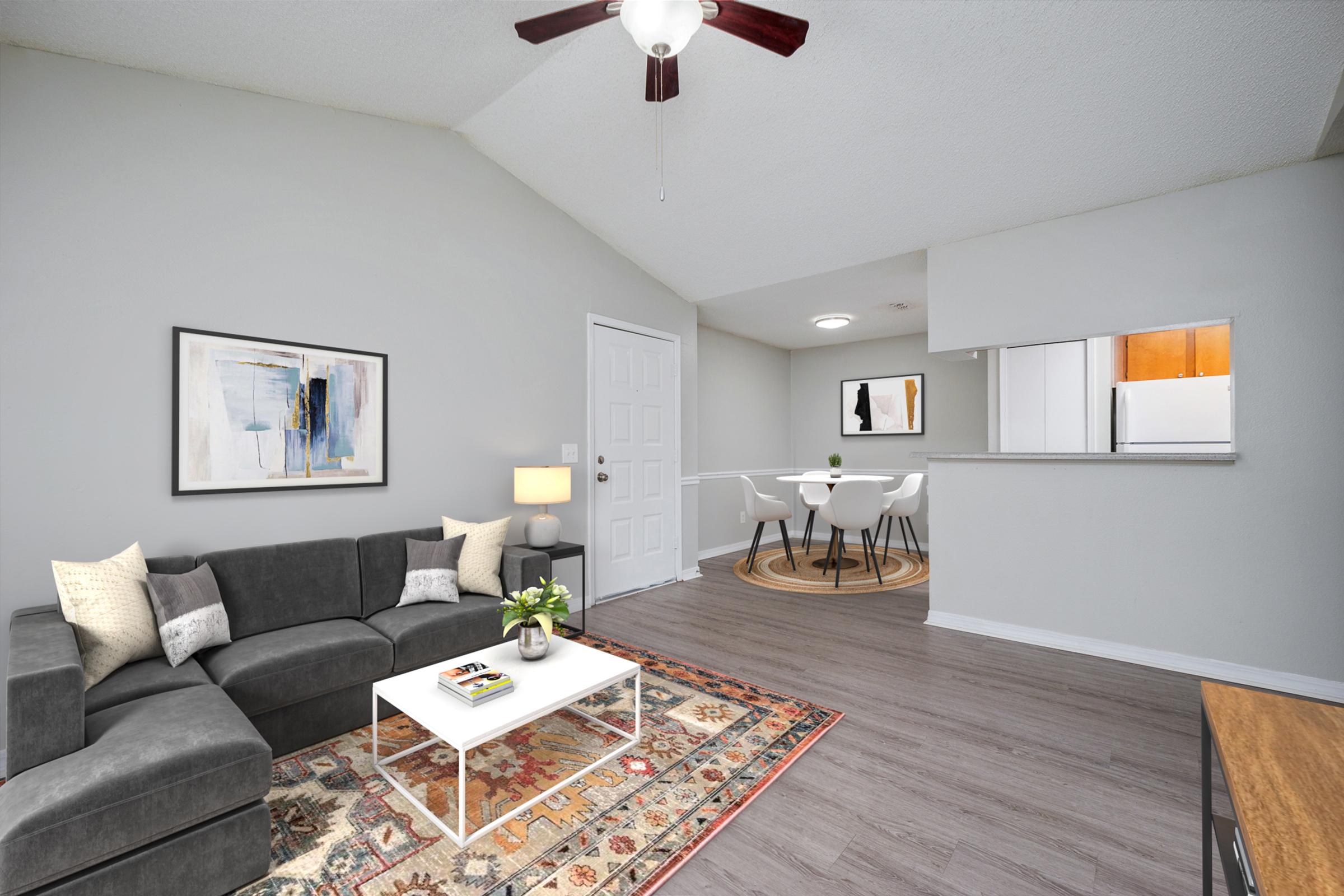 A modern, well-lit living room featuring a gray sectional sofa, white coffee table, and a small decorative plant. A colorful area rug adds warmth to the space. The walls are painted light gray, and there's a dining area with a round table and white chairs visible in the background, along with a kitchen area.