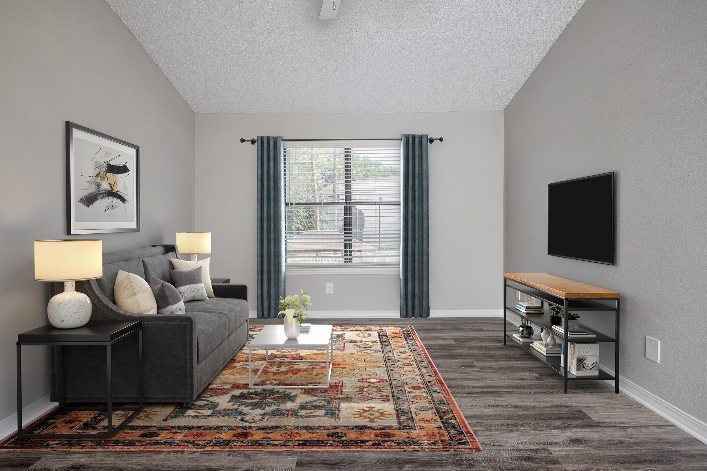 A modern living room featuring a gray sofa, two lamps, and a wooden coffee table. A large area rug with intricate patterns is on the floor. There are large windows with sheer curtains allowing natural light, and a minimalist TV stand against the wall, enhancing the cozy atmosphere.