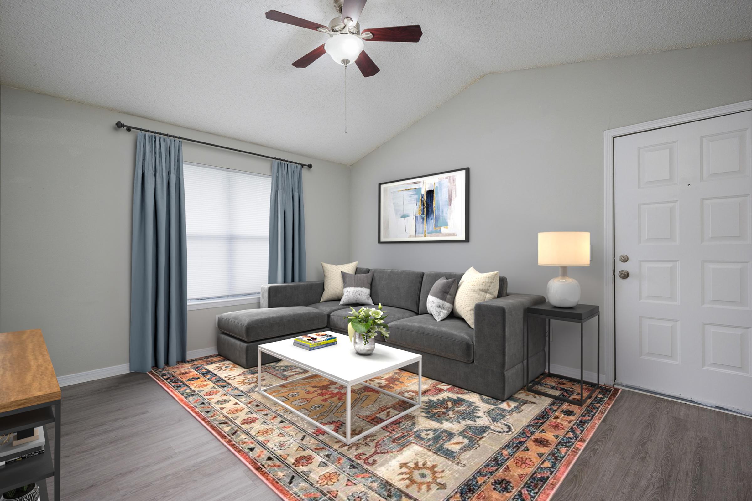 A cozy living room featuring a gray sectional sofa with decorative pillows, a white coffee table on a colorful area rug, and a lamp beside the entrance. The walls are light gray, with a window adorned by blue curtains, creating a bright and inviting atmosphere. A piece of artwork hangs above the sofa.