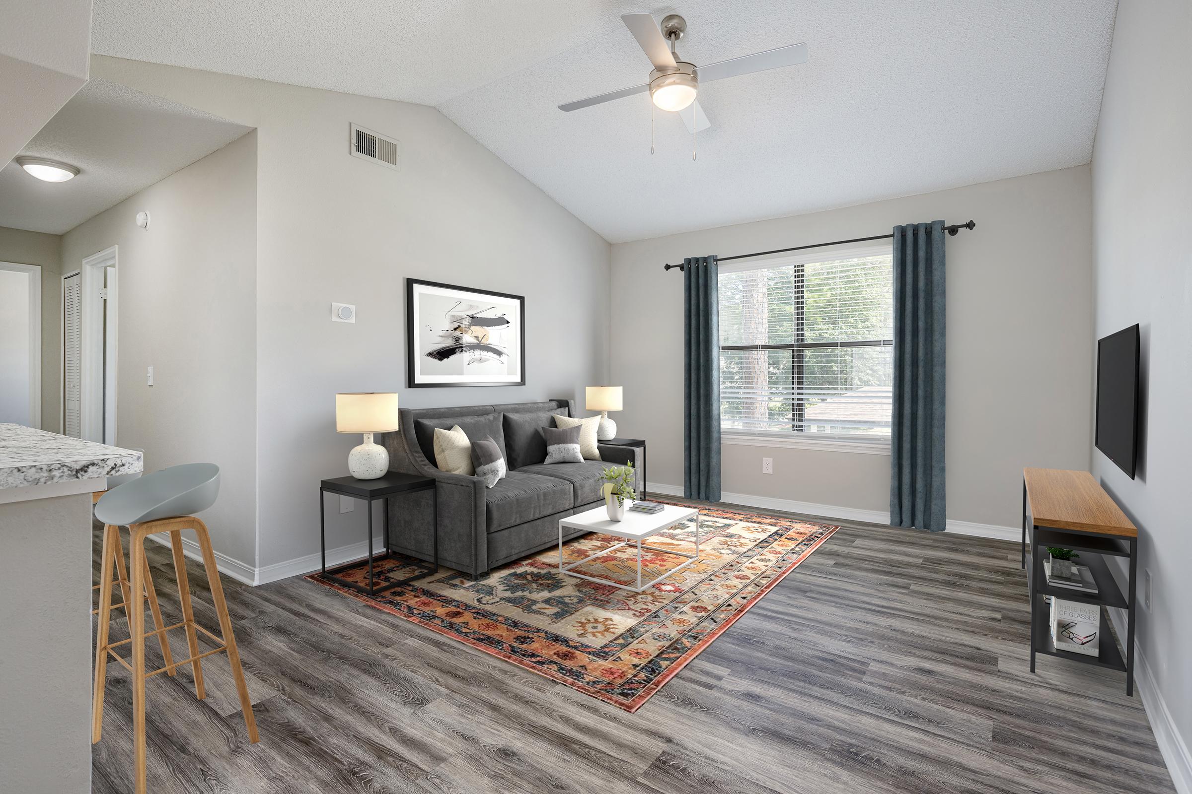 A modern living room featuring a gray sofa with two matching lamps on either side, a coffee table with a small plant, and a colorful area rug. There are two large windows with blue curtains letting in natural light, and a mounted television on a wooden stand. The walls are painted in a light color, enhancing the open feel.
