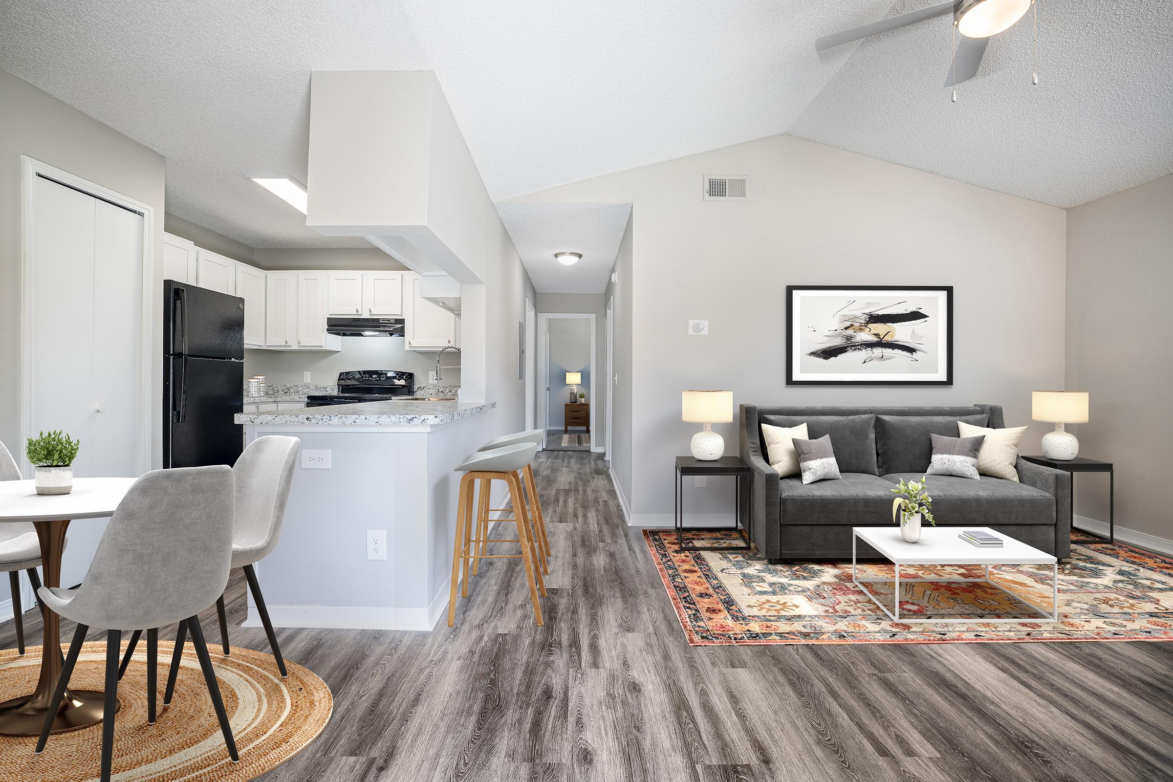 Modern living room and kitchen area with a cozy gray sofa, decorative artwork on the wall, and a circular rug. Light wood flooring and a small dining table with chairs. Kitchen features white cabinets and a black refrigerator. Brightly lit space with ceiling fan, showcasing an open and inviting layout.