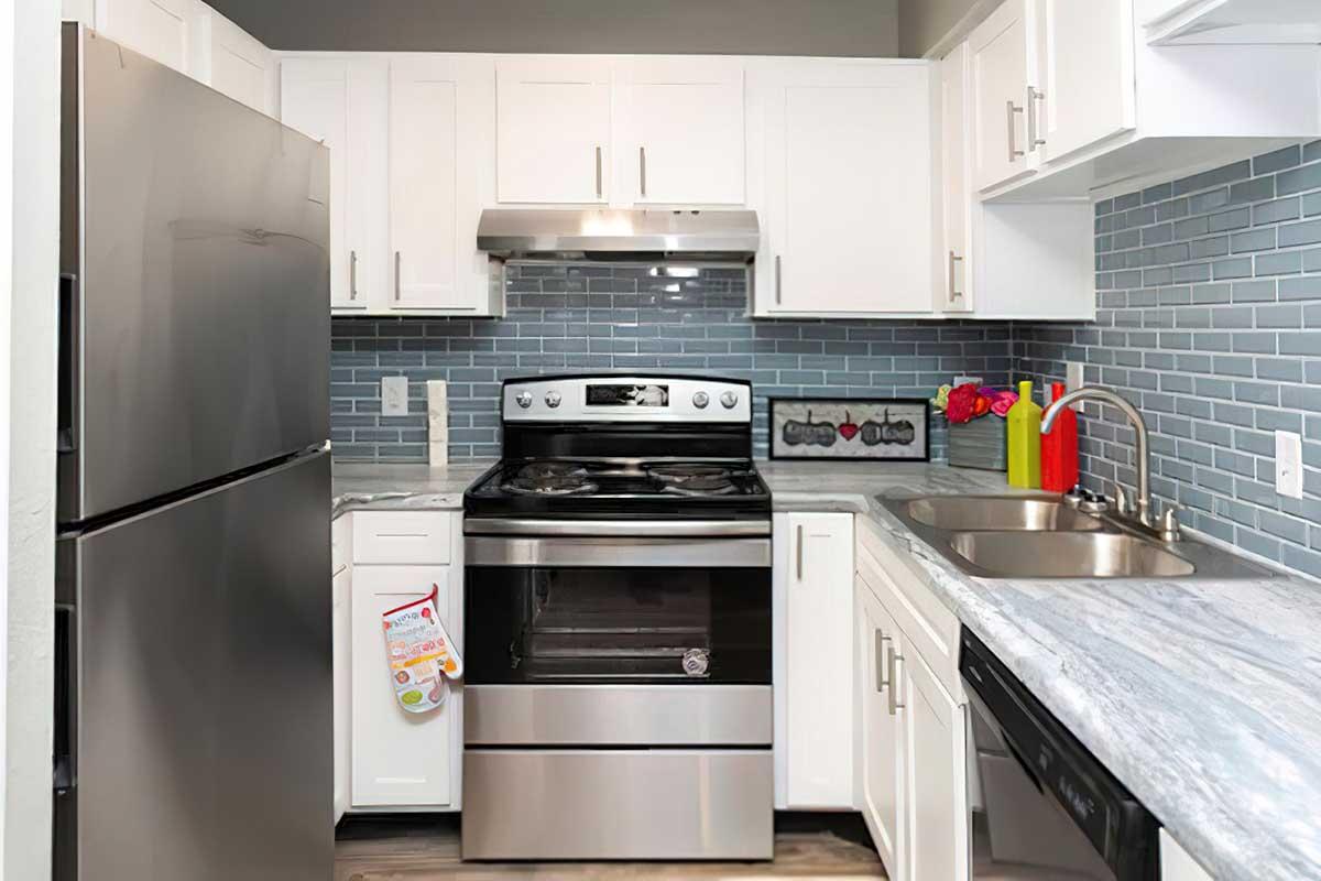 Modern kitchen featuring stainless steel appliances, including a refrigerator and an oven with a stovetop. The cabinetry is white with a marble-like countertop. A decorative backsplash in shades of blue complements the kitchen's aesthetic, while a sink and red flowers add a pop of color.