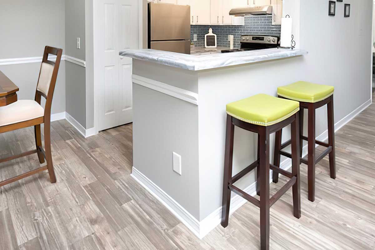 A modern kitchen interior featuring a marble-topped island with two green cushioned bar stools. The room has light gray walls and wooden flooring, with a dining table and chairs visible in the background, along with a stainless steel refrigerator and stove.