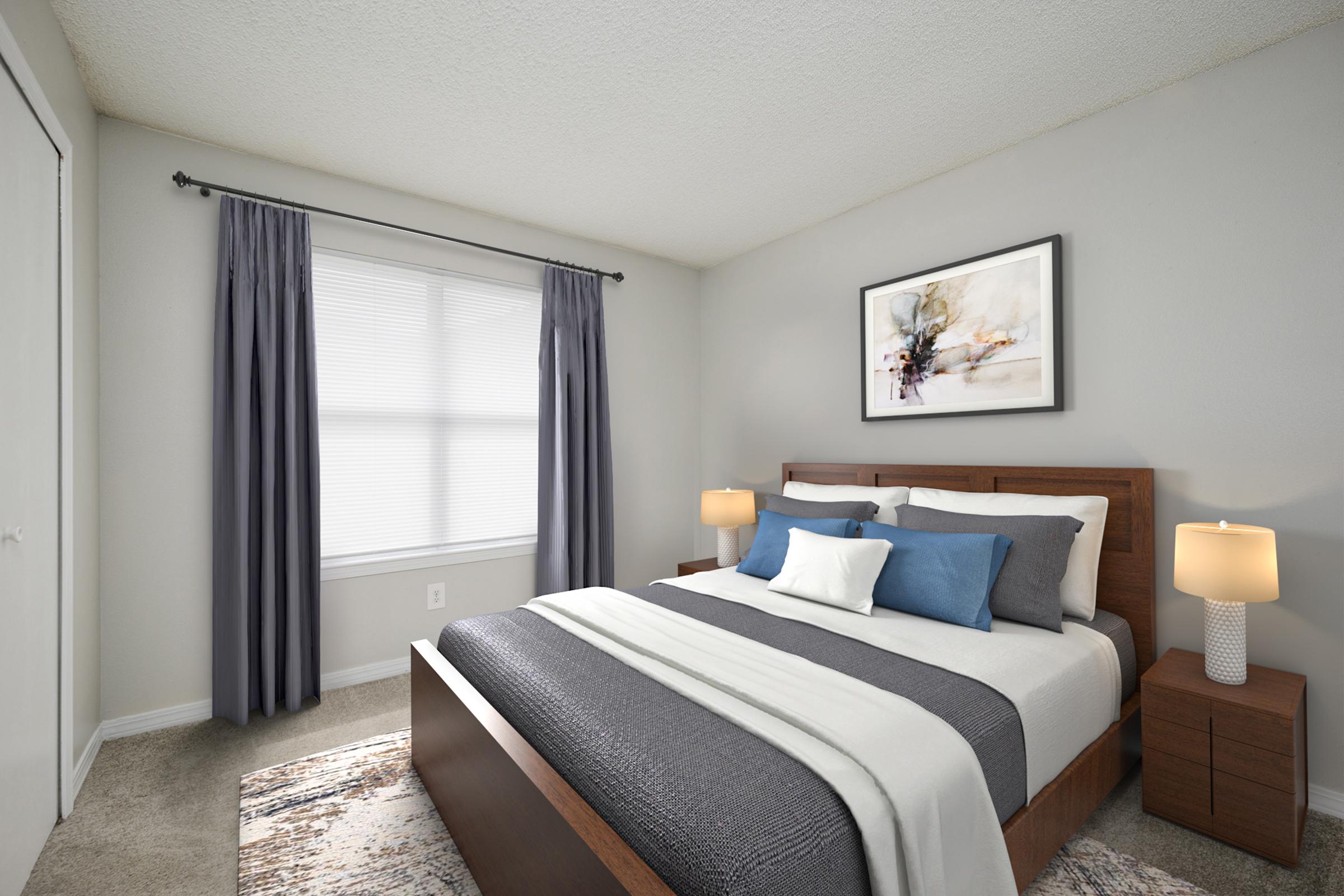 A cozy bedroom featuring a wooden bed with gray and blue bedding, two bedside tables with lamps, a large window with sheer curtains, and an abstract painting on the wall. The walls are light gray, and the room has a soft, neutral carpet, creating a warm and inviting atmosphere.