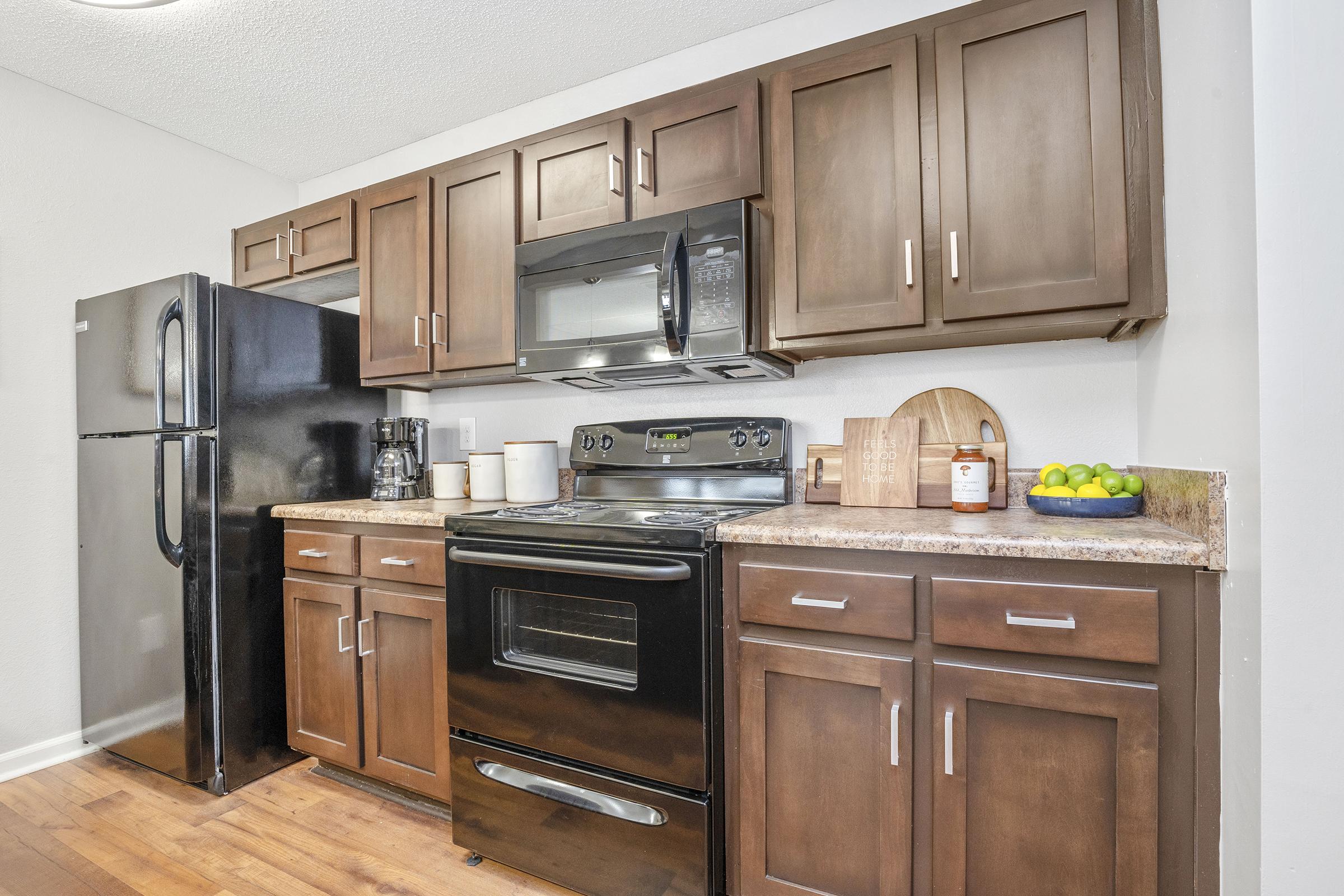 Modern kitchen featuring dark wooden cabinets, a black refrigerator, a microwave above the stove, and a granite countertop. Kitchen utensils and a bowl of citrus fruits are neatly arranged on the counter, with warm wooden flooring enhancing the inviting atmosphere.