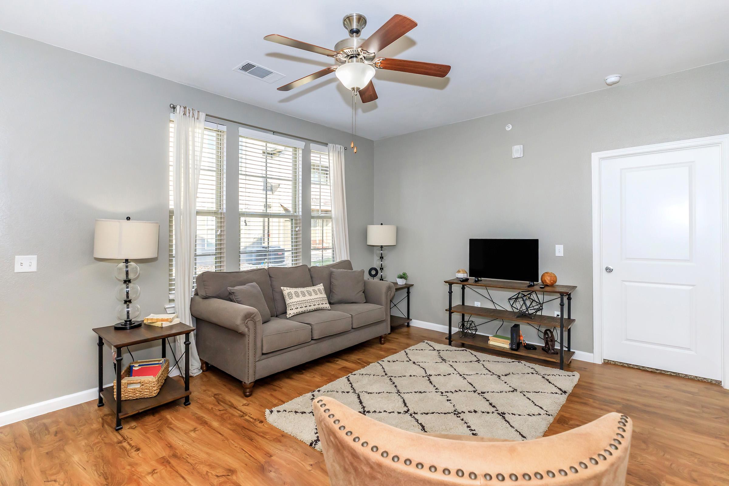 A cozy living room featuring a gray sofa, two side tables with lamps, a coffee table with a decorative item, and a TV on a stand. Natural light floods the space through large windows with blinds. The floor is wood, and there's a patterned area rug beneath the coffee table.