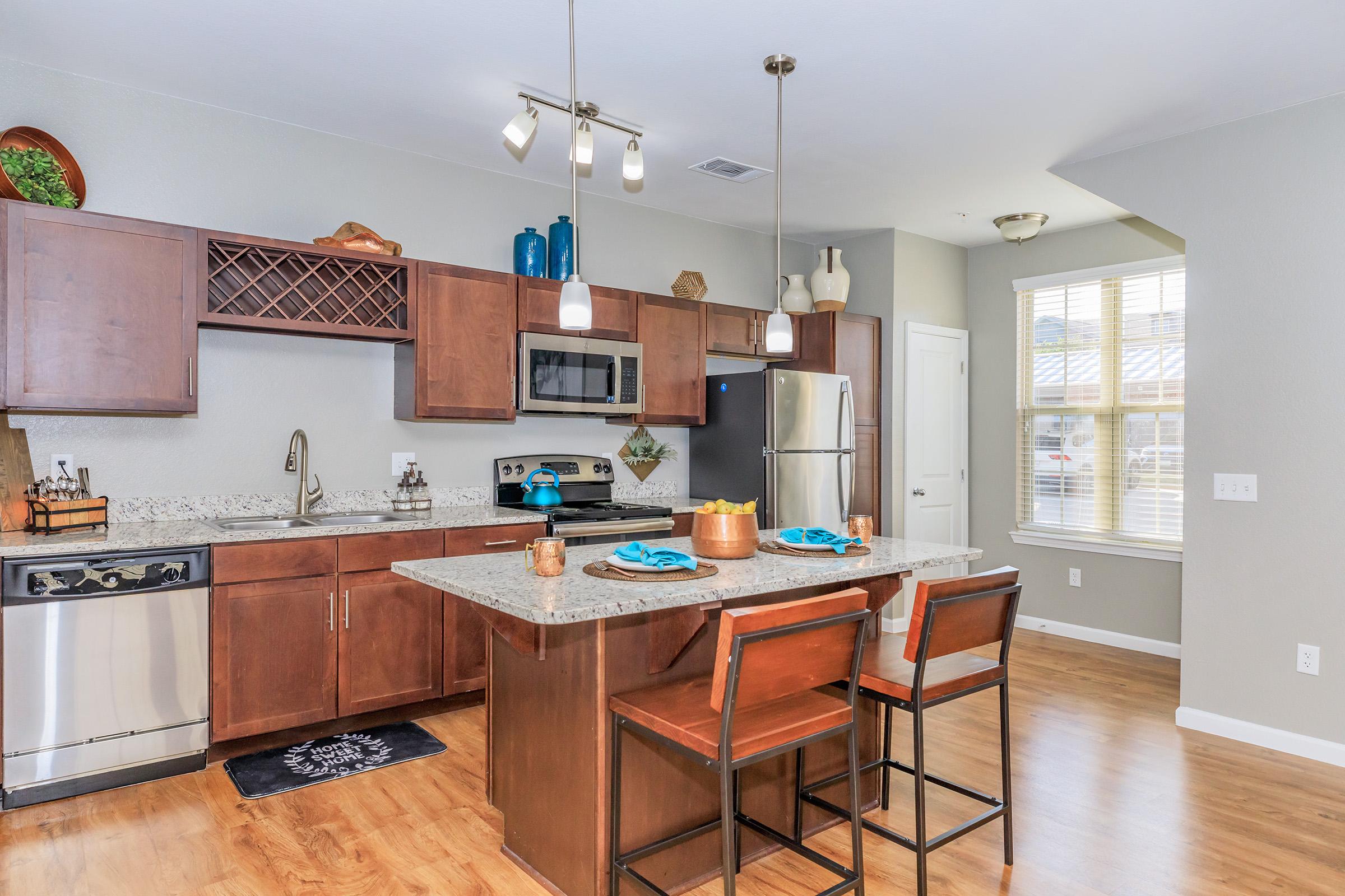 Modern kitchen featuring wooden cabinetry, granite countertops, stainless steel appliances including a microwave and dishwasher, and a center island with seating for two. There are decorative items on the shelves, and large windows allowing natural light to fill the space.
