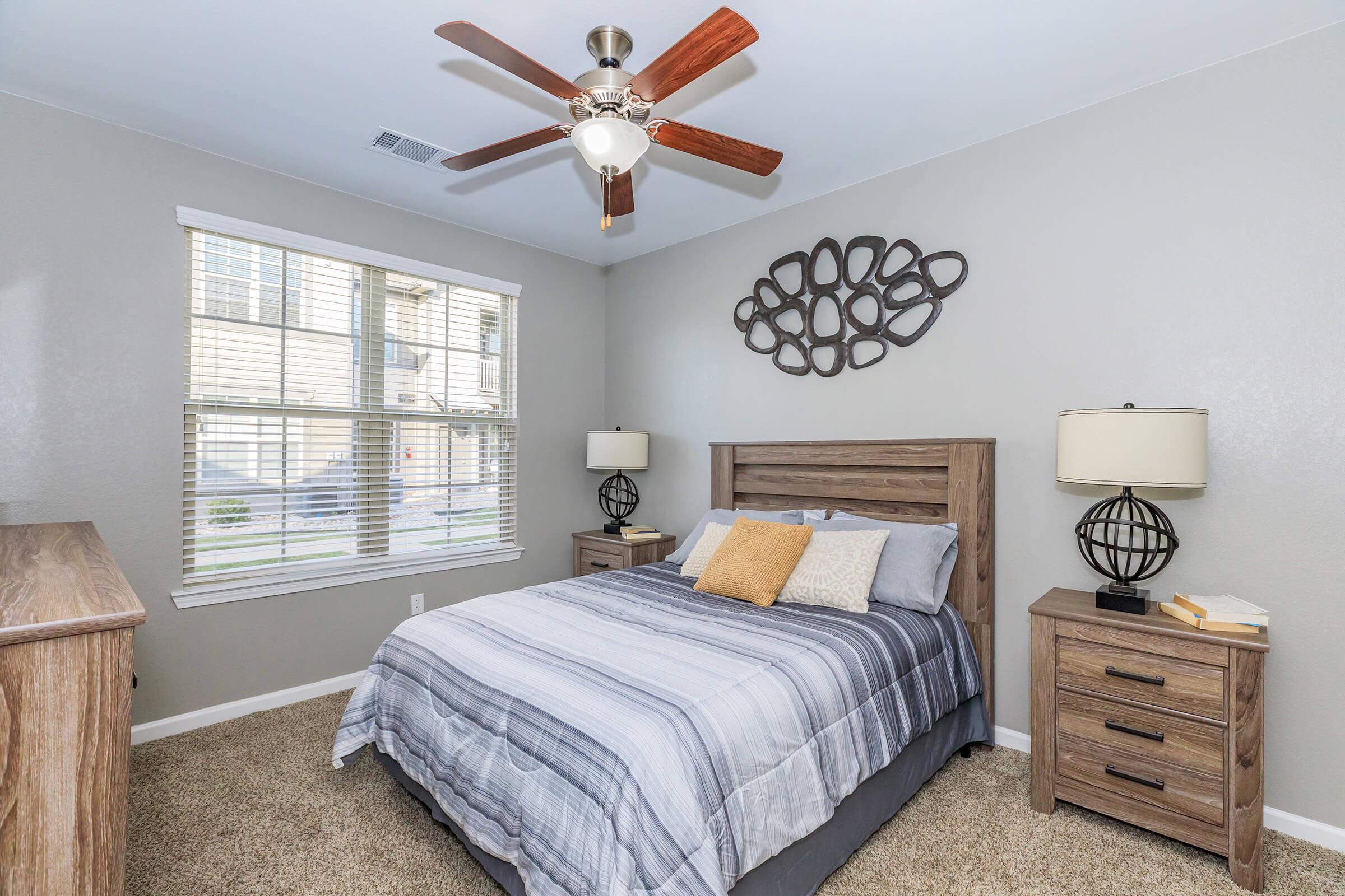 Cozy bedroom featuring a queen-sized bed with gray and white striped bedding, two bedside tables with decorative lamps, a wooden dresser, and a modern ceiling fan. Natural light streams in through a window adorned with blinds, and a unique wall decoration adds a touch of style to the room.