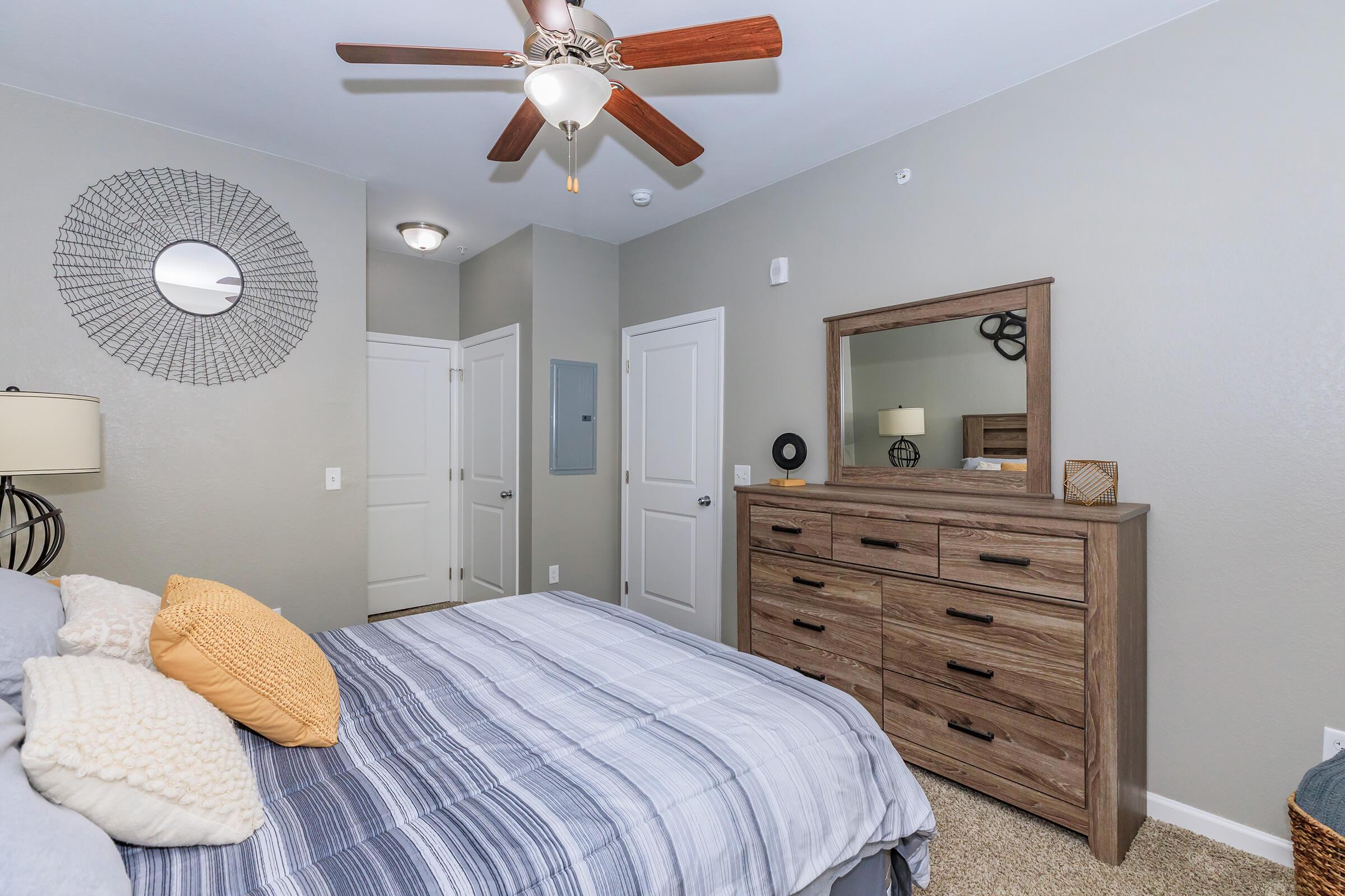 A cozy bedroom featuring a comfortable bed with gray and blue striped bedding, an orange accent pillow, a wooden dresser, a round wall mirror, and a ceiling fan. Light walls and carpet create a warm, inviting atmosphere. A door leads to another room, enhancing the room's spacious feel.