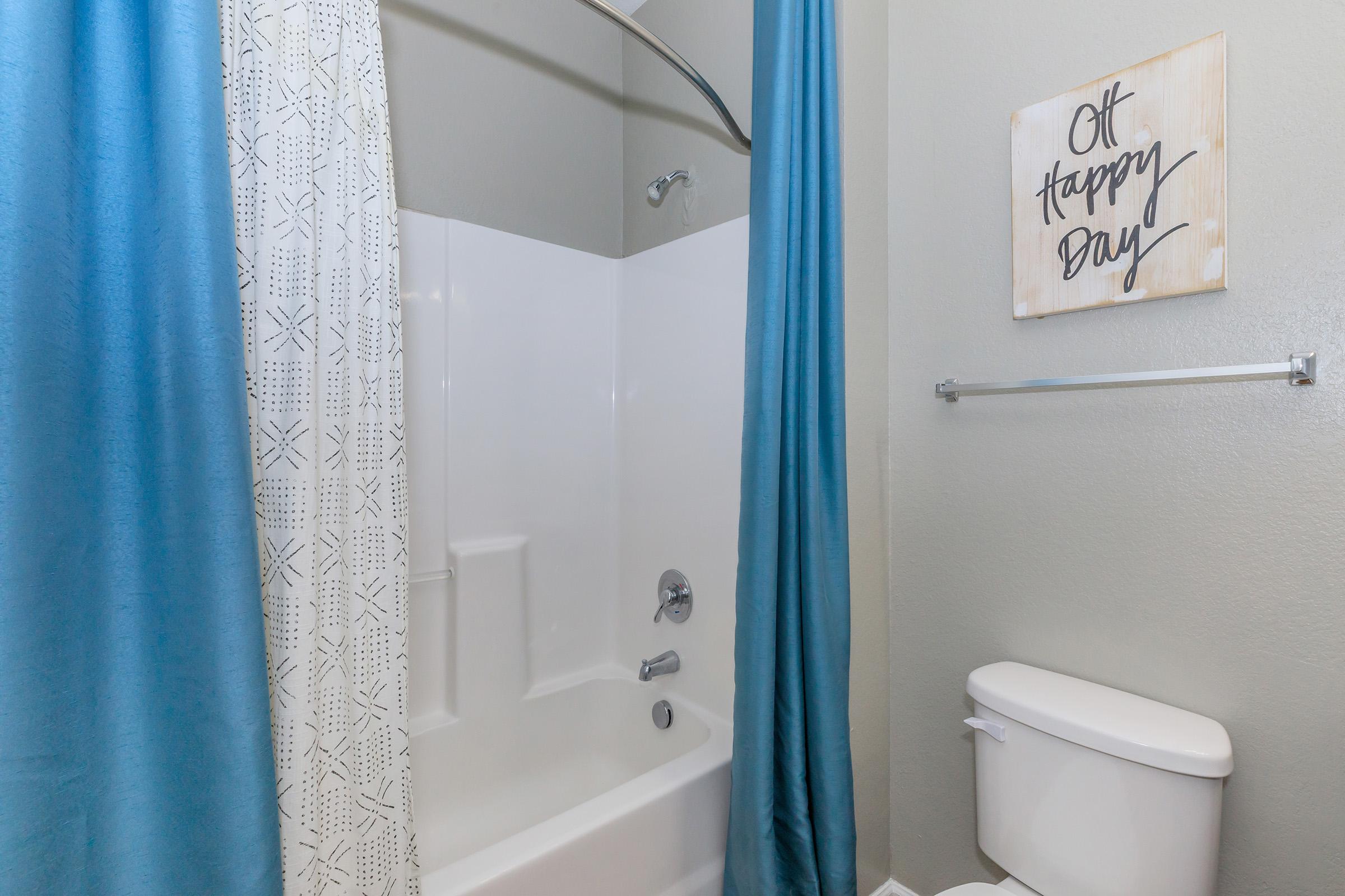 A modern bathroom featuring a white bathtub with a shower curtain in blue and patterned fabric. The wall is painted light gray, and a framed sign reading "Oh Happy Day" is hanging nearby. A white toilet is visible next to the tub, completing the clean and cheerful aesthetic of the space.