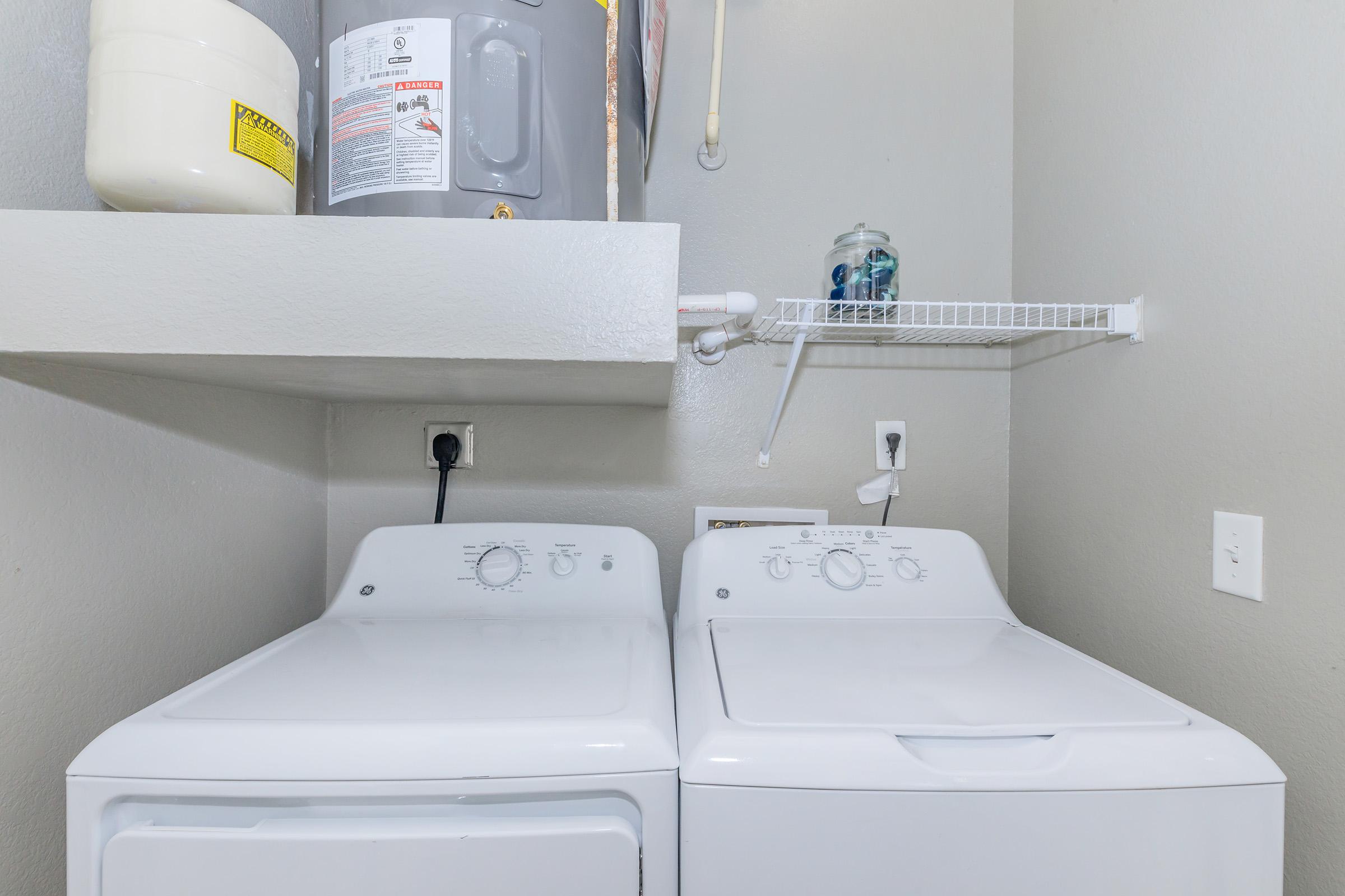 A laundry room featuring a top-loading washing machine and a dryer side by side. Above them, there is a shelf with a water heater and a glass container. The wall is painted in a light gray color, and there are electrical outlets and a pipe visible. The space is organized and functional.