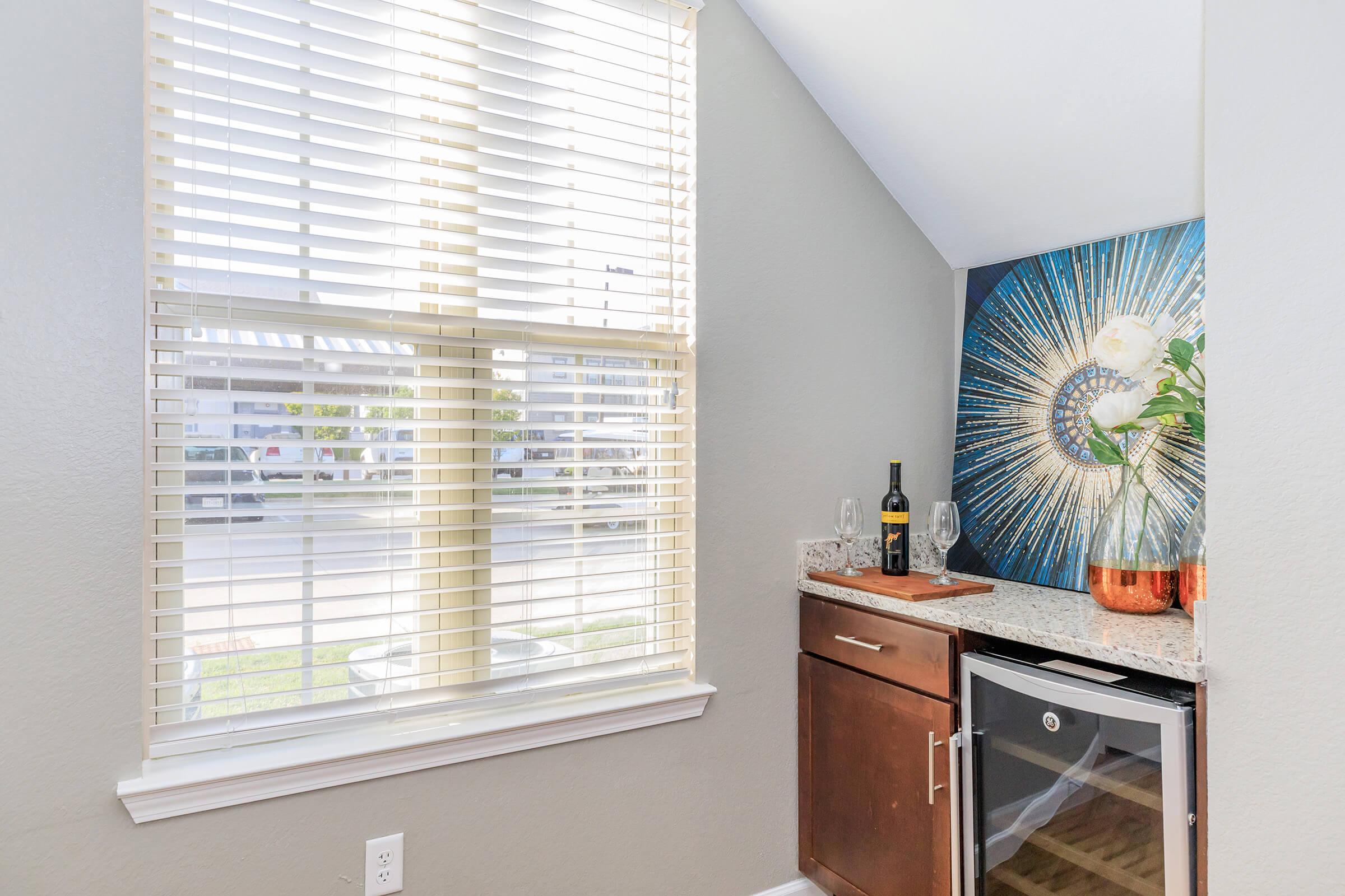 Bright corner of a kitchen featuring a large window with blinds, letting in natural light. A decorative wall art piece with a sunburst design hangs above a countertop with a wine bottle, two glasses, and a wine cooler. The scene conveys a modern and inviting atmosphere.