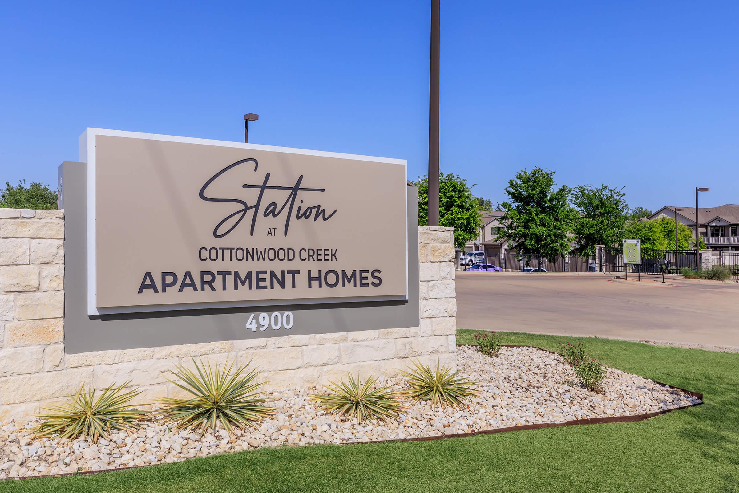 Sign for "Station at Cottonwood Creek Apartment Homes" featuring modern design, located at 4900. The surrounding area includes landscaped greenery and clear blue skies, with a driveway and buildings visible in the background.