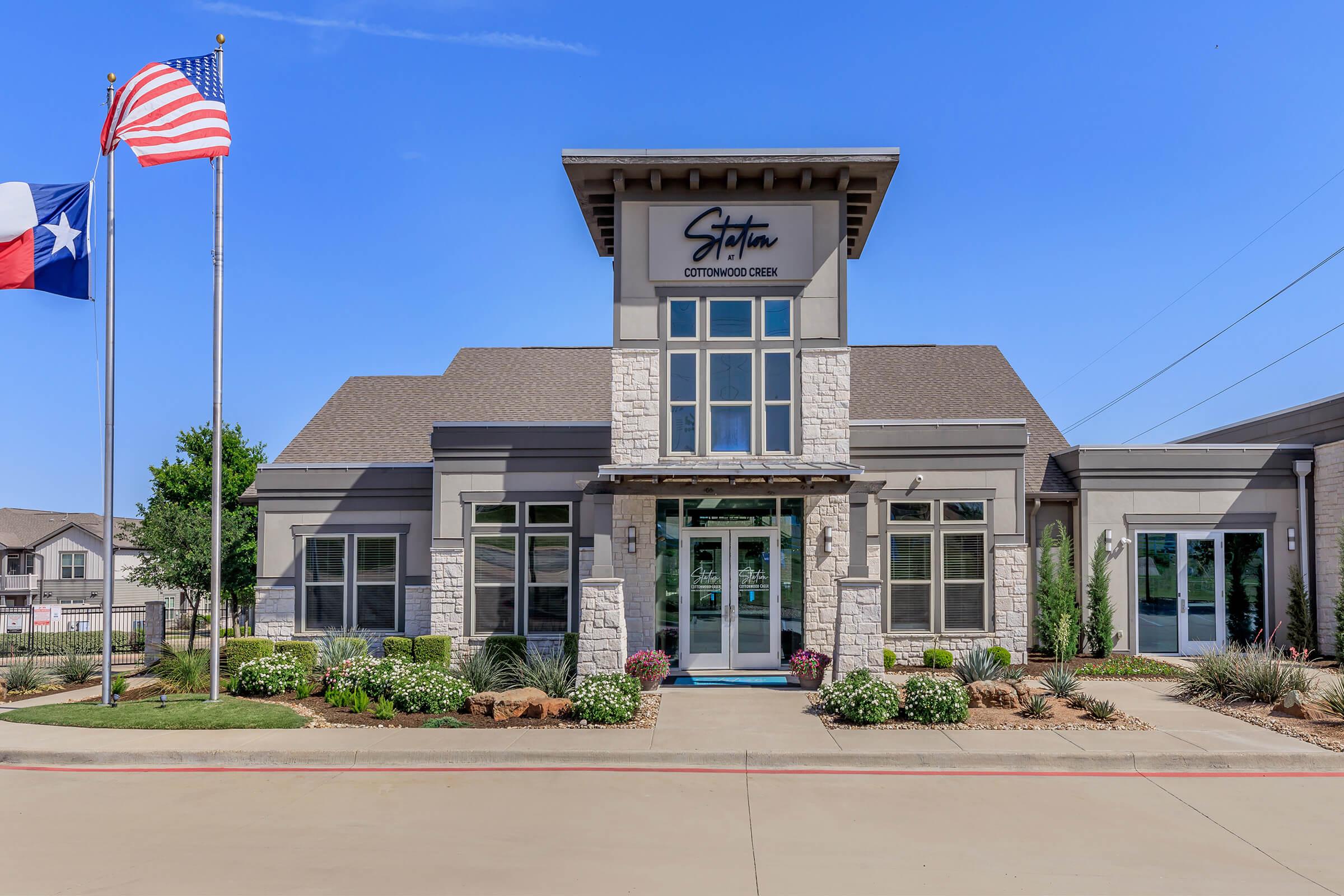A modern building with a stone and siding facade, featuring large windows and a welcoming entrance. Flags of the United States and Texas are displayed in front. The landscape includes neatly arranged plants and a well-maintained pathway, set against a clear blue sky.