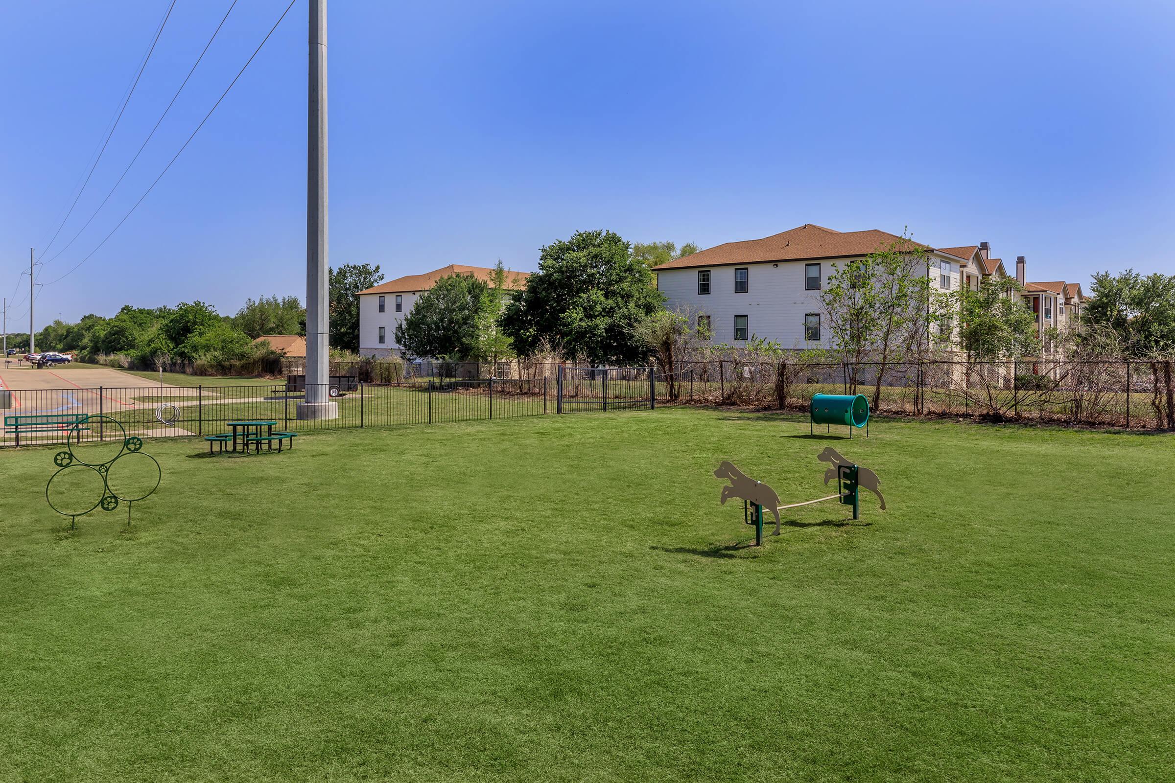 A grassy park with a playground area featuring metal sculptures of animals, a green cylindrical object, and picnic tables. In the background, there are residential buildings and utility poles under a clear blue sky.