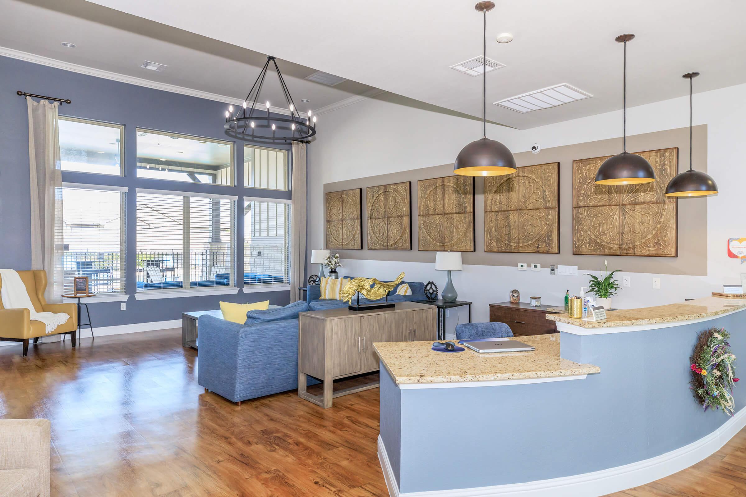 A modern interior of a living space featuring a blue couch, a wooden coffee table, and a yellow decorative piece. There are large windows letting in light, a chandelier hanging from the ceiling, and wall art made of four textured panels. A small workspace with a desk is visible.
