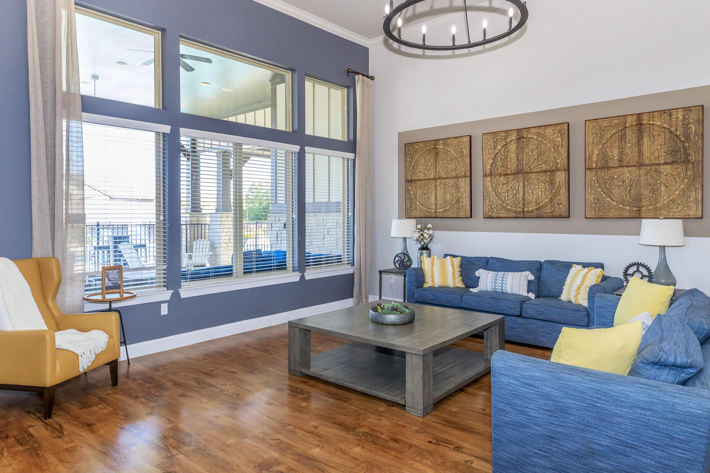 A bright and spacious living room featuring large windows with natural light, a cozy blue sofa with yellow accents, a wooden coffee table, and decorative wall art. A stylish armchair adds to the decor, with neutral paint colors and wood flooring enhancing the inviting atmosphere.