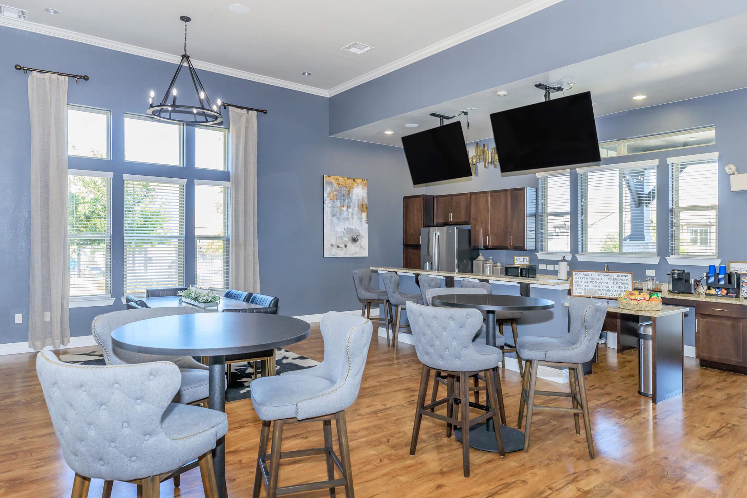 A modern community lounge featuring blue-gray walls, large windows with natural light, stylish furniture including round tables and tufted chairs, a kitchenette with dark wood cabinets, and two mounted flat-screen TVs. The space is inviting and designed for social gatherings.