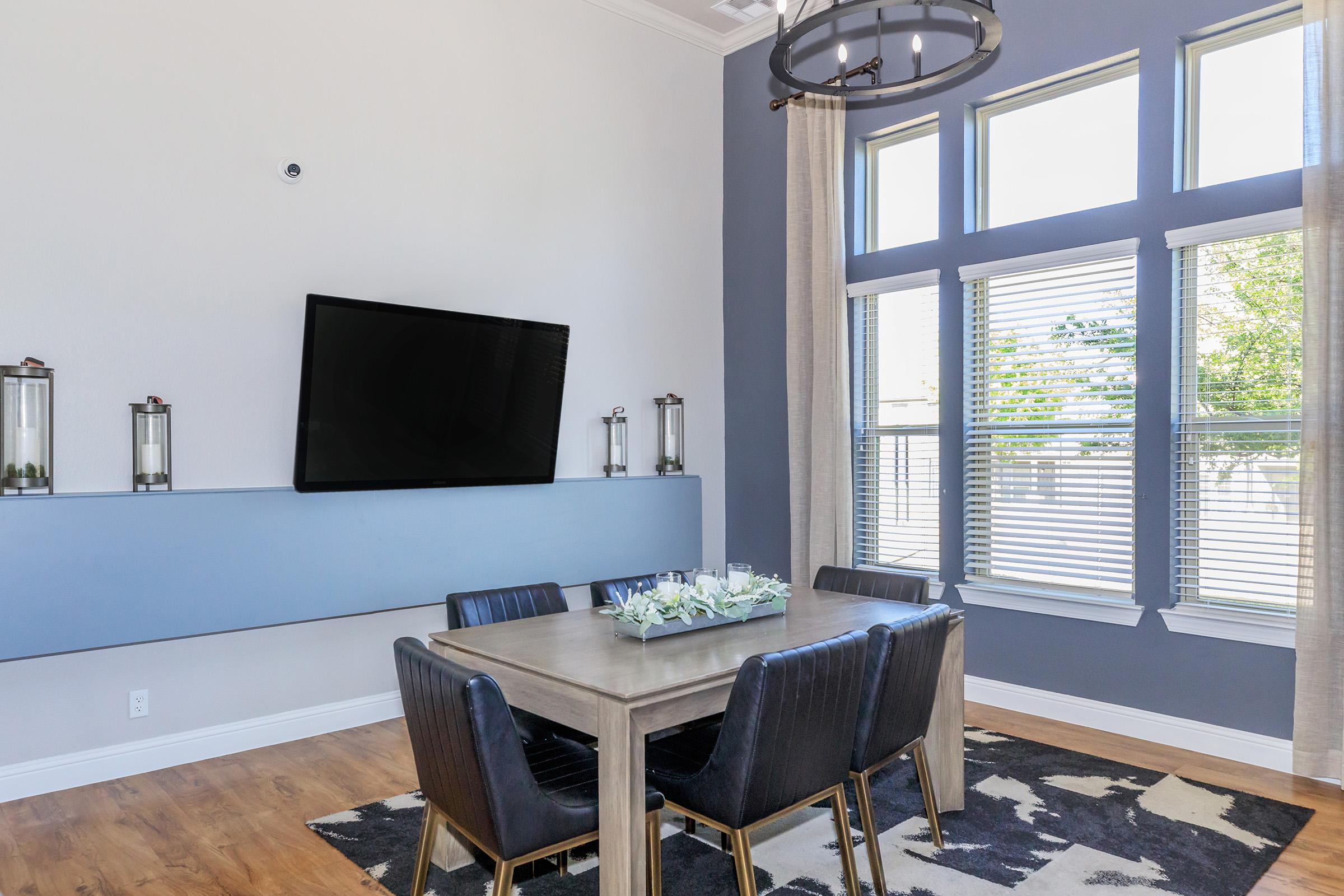 A modern dining area featuring a rectangular wooden table with dark leather chairs. A large flat-screen TV mounted on a light blue wall is above a decorative console. Bright sunlight streams through tall windows with blinds, and a patterned rug is under the table, enhancing the stylish decor.