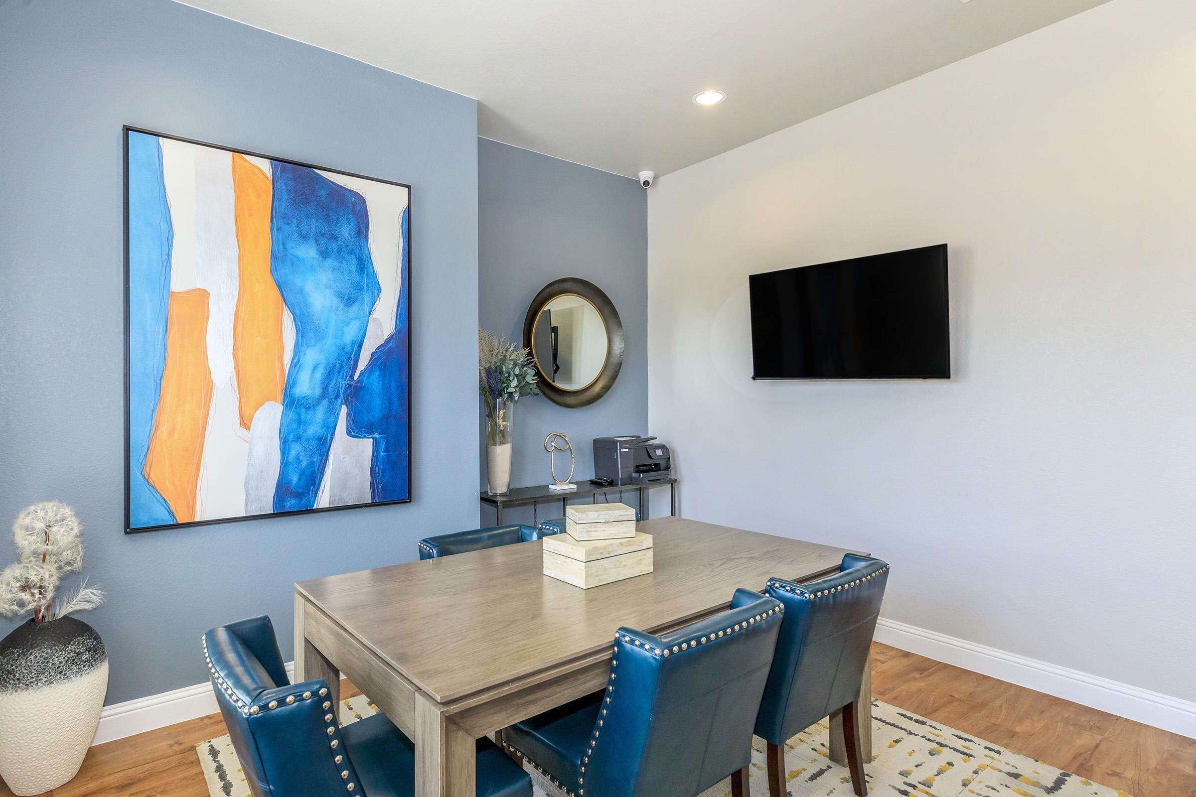 A modern dining area featuring a wooden table surrounded by blue chairs, with an abstract painting on the wall. A round mirror enhances the space, and a television is mounted on the wall. Decorative elements include a vase and a light-colored rug on the floor.