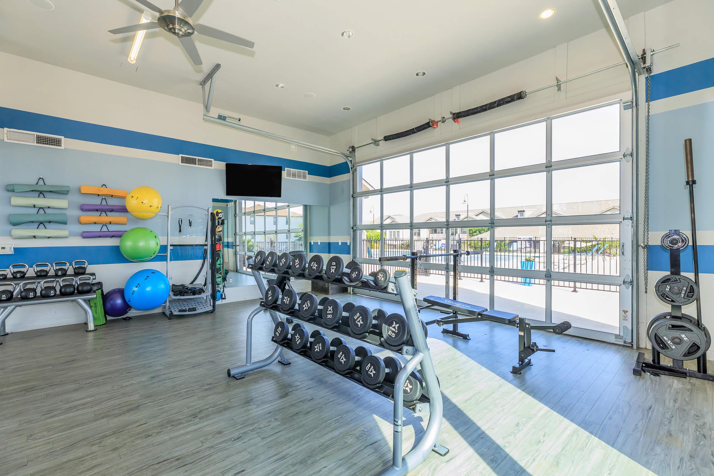A bright and well-equipped fitness room featuring dumbbells, exercise balls, a bench, and a large window with a view. The space is modern, with light wood flooring and blue accents, creating an inviting atmosphere for workouts.
