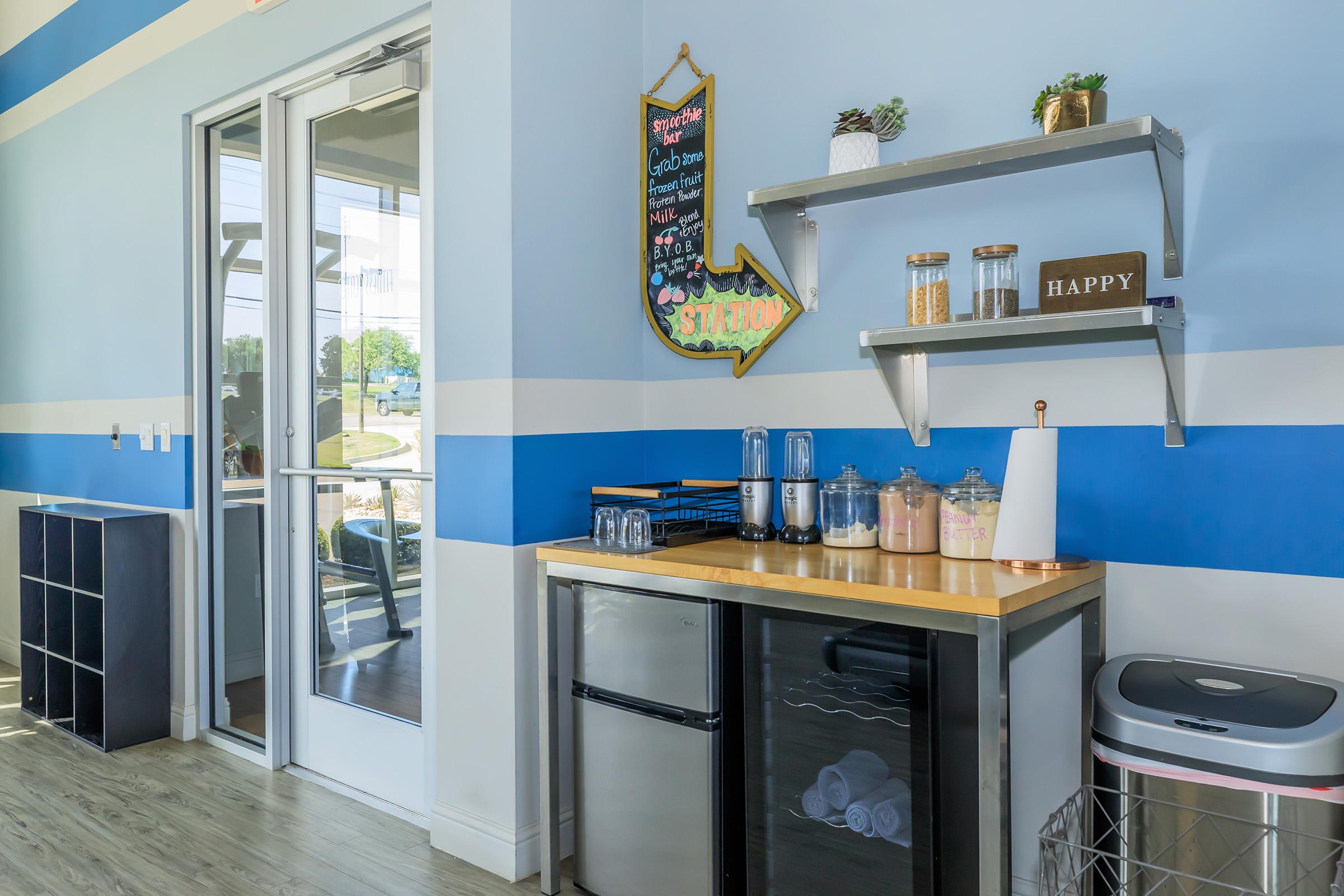 A modern kitchen corner featuring a small fridge and countertop with blenders, containers, and decorative items. A wall shelf displays jars and the word "HAPPY." Large glass doors lead outside, and the walls are painted in blue and white stripes. The space is bright and organized.