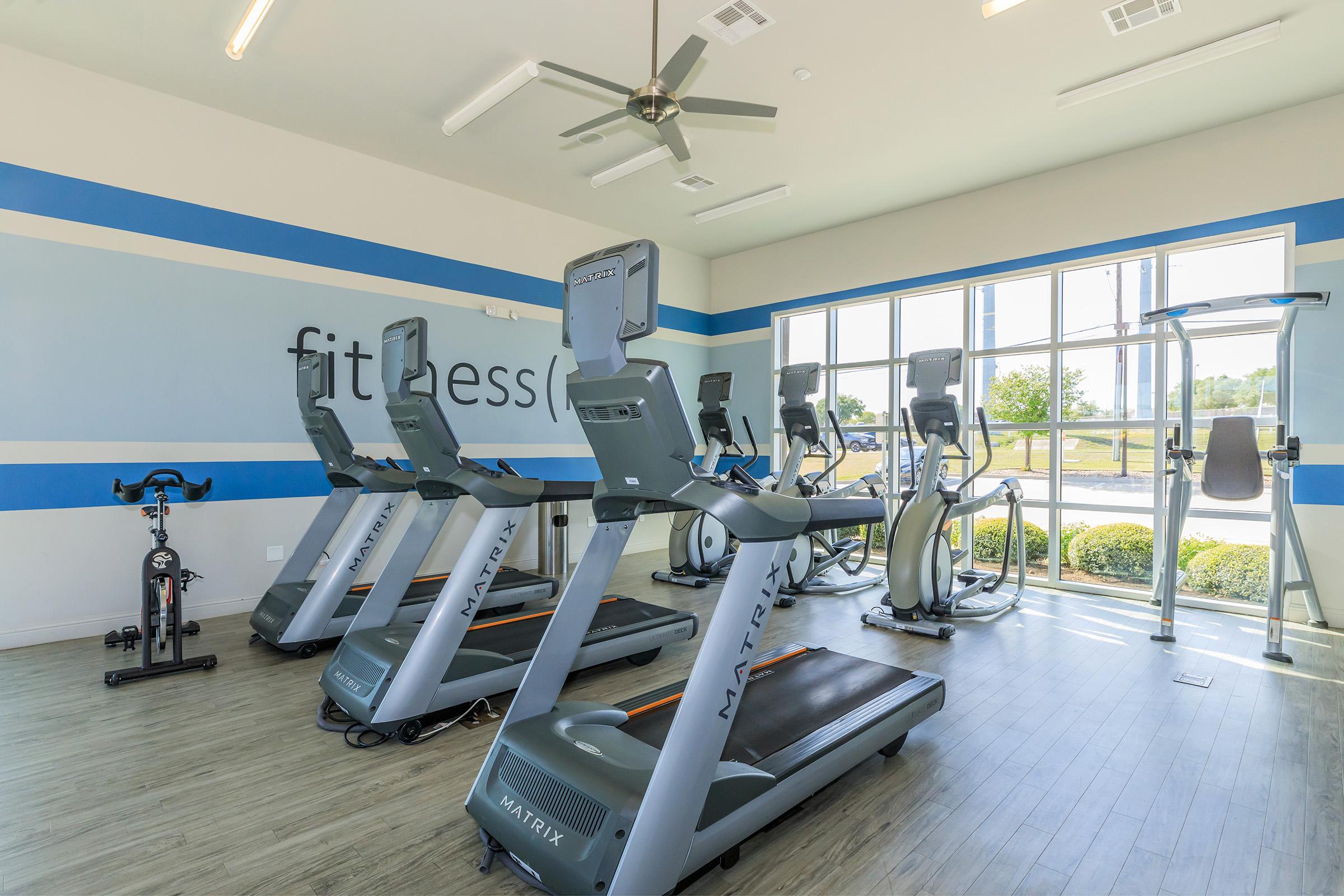 A modern fitness room featuring several Matrix exercise machines, including treadmills and stationary bikes. Large windows allow natural light to fill the space, with a blue and white color scheme on the walls. The floor is composed of light wood, enhancing the bright and inviting atmosphere.