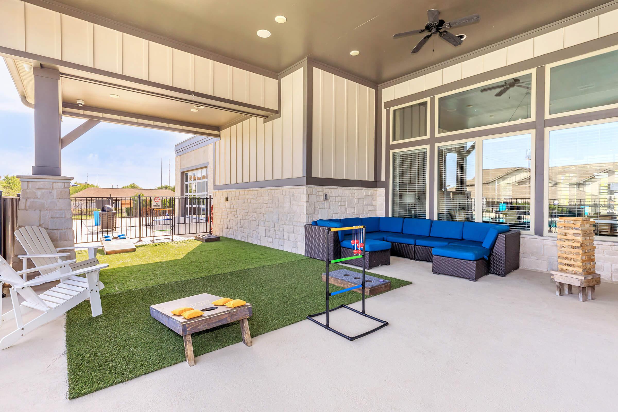 Outdoor patio area featuring a green turf space, a blue sectional sofa, and a small coffee table. There is a game setup on a stand and a few games like cornhole on the ground. Sunlight illuminates the space, which is surrounded by a decorative fence and large windows.