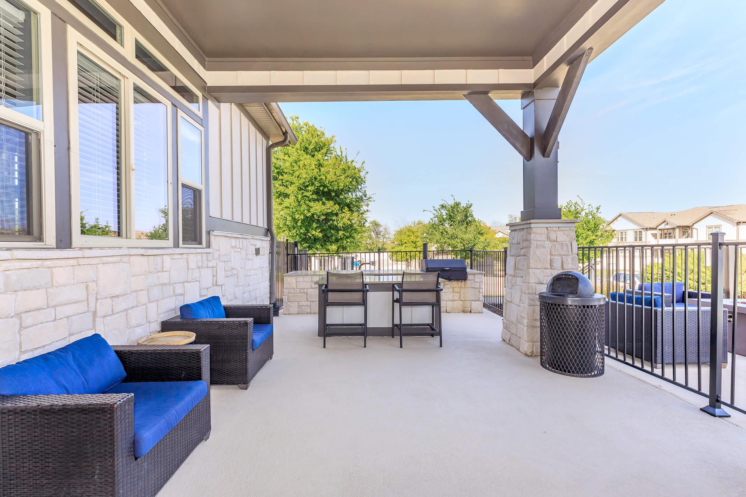 Patio area featuring two blue cushioned chairs and a small table, surrounded by stone pillars. There is a grill in the background, a trash can, and a view of greenery beyond the railing. Bright blue sky overhead, creating a welcoming outdoor space for relaxation.