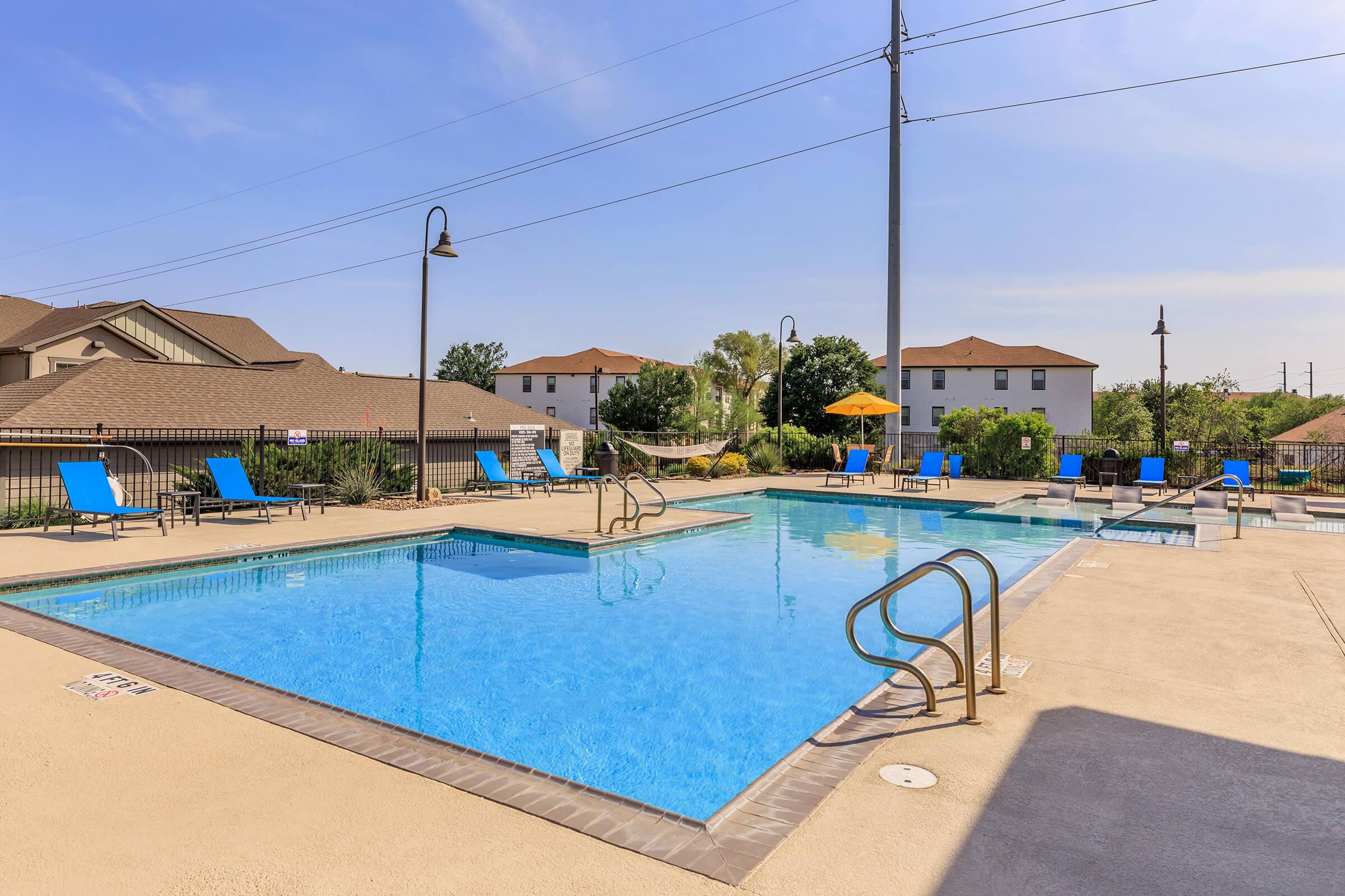 A clear swimming pool surrounded by lounge chairs and umbrellas on a sunny day. In the background, residential buildings and trees are visible. The pool area features a light concrete deck and is equipped with safety rails.