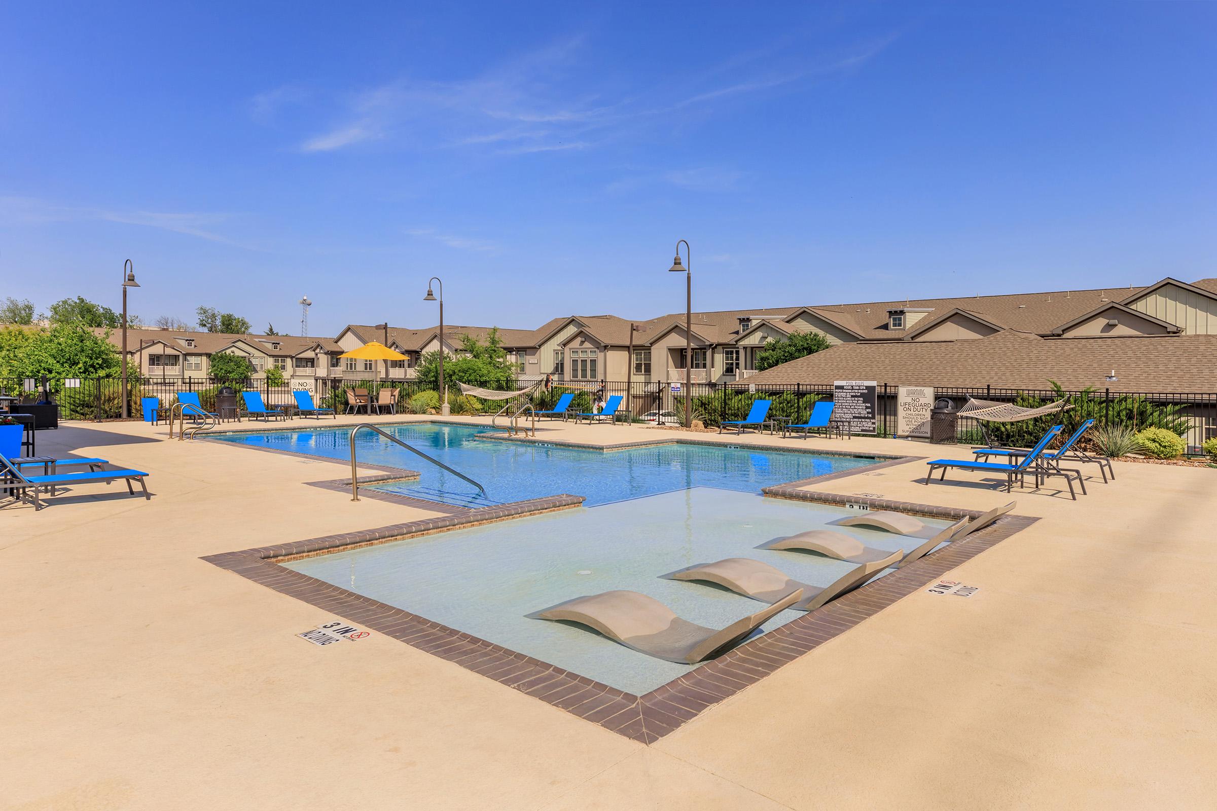 A bright outdoor swimming pool area featuring a rectangular pool with shallow lounging areas, surrounded by blue lounge chairs. Residential buildings are visible in the background under a clear blue sky. The setting appears inviting and perfect for relaxation.