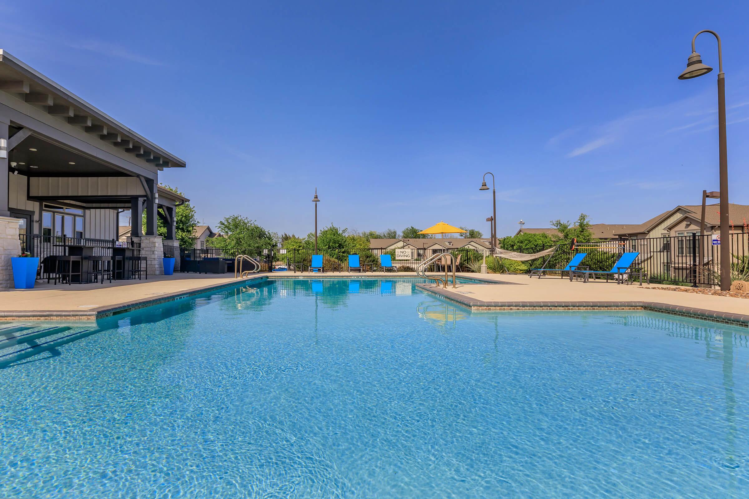 A serene swimming pool surrounded by a patio with lounge chairs, set against a clear blue sky. Nearby, there is a shaded area with seating, and lush greenery borders the pool area, creating a relaxing outdoor space.