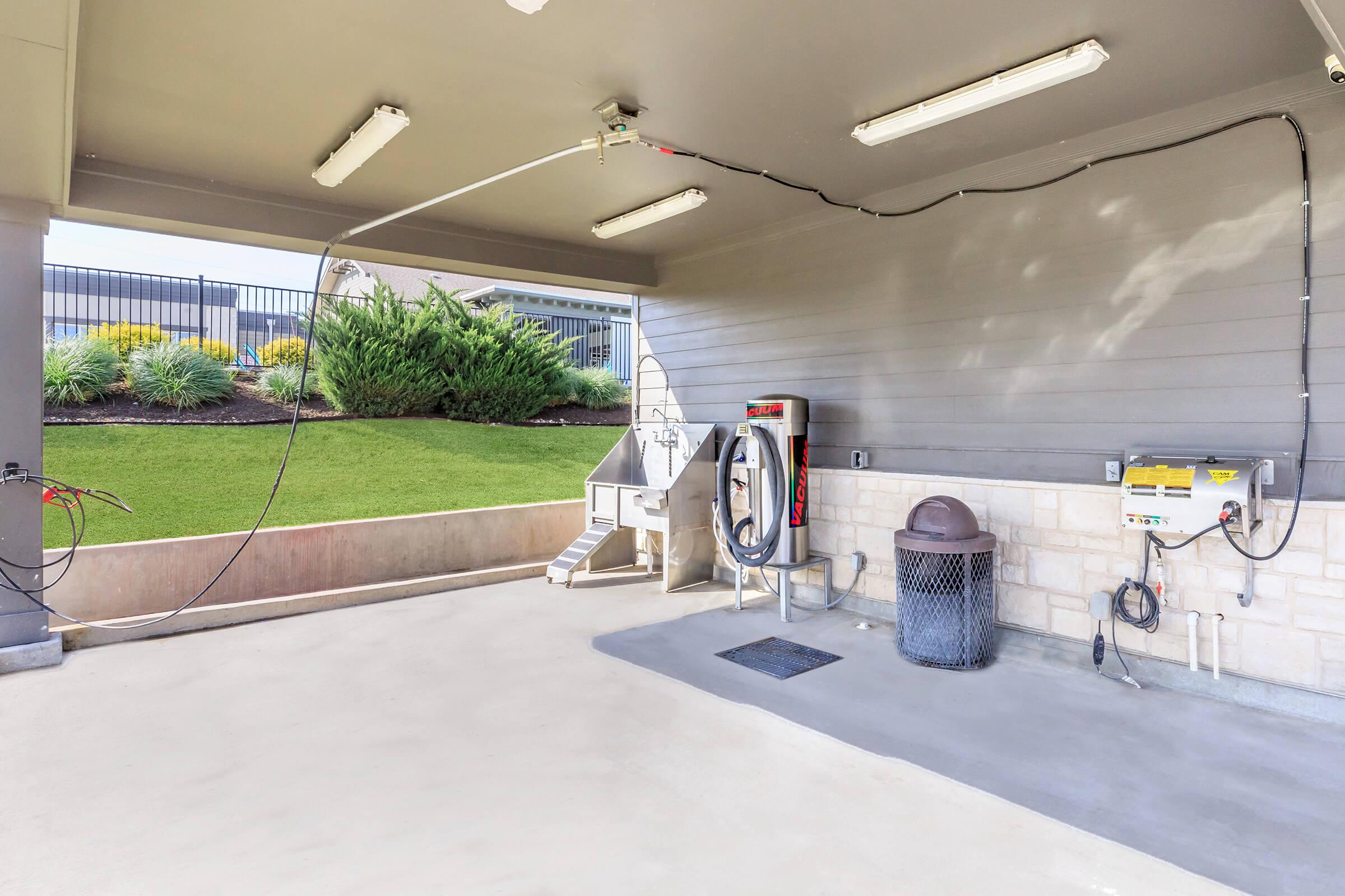 A clean and spacious covered area featuring a washing station with a hose and equipment, a trash bin, and a grassy landscape visible outside. The setting has bright fluorescent lights, a concrete floor, and modern fixtures, creating a functional and tidy environment.