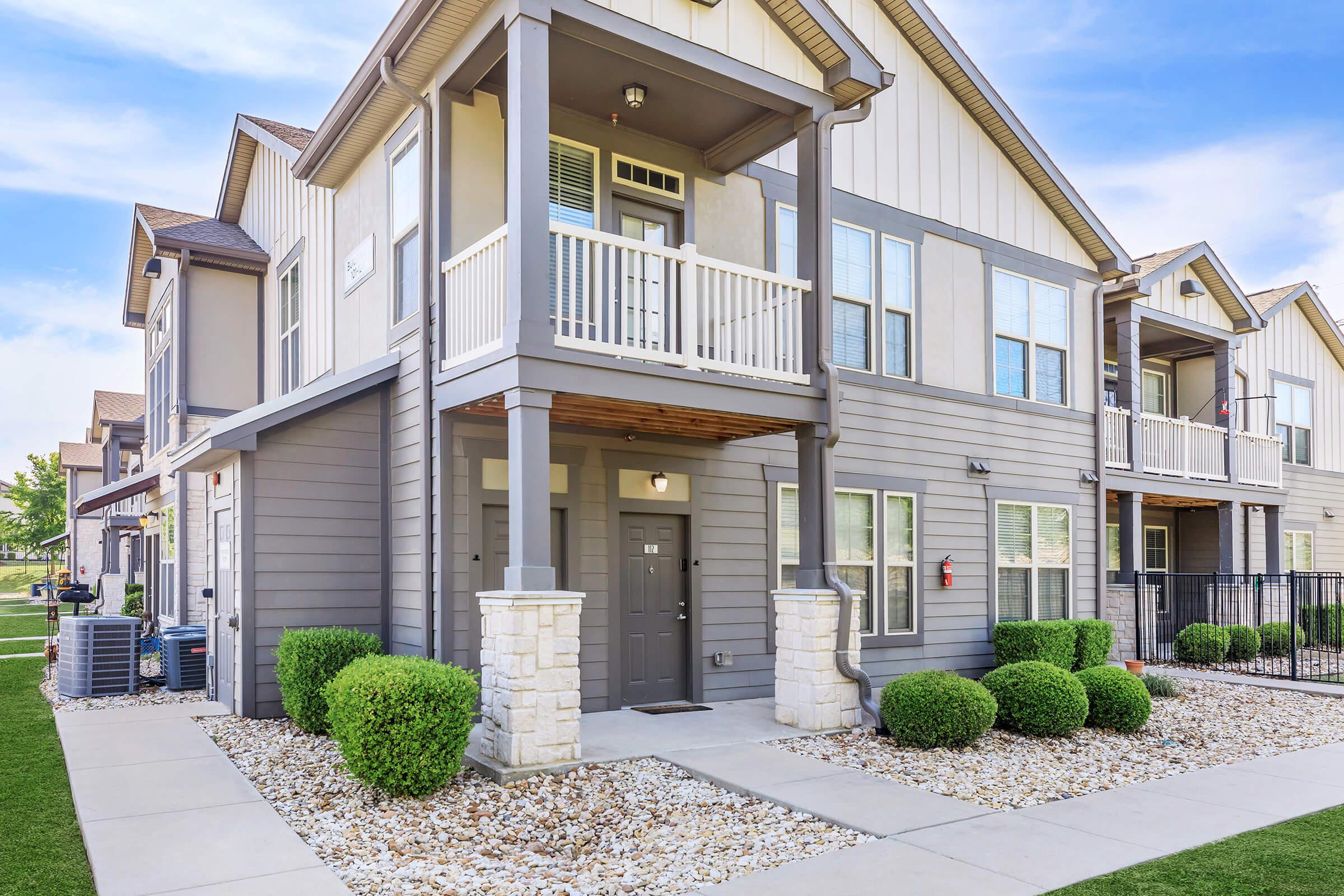 A modern apartment building features a gray exterior with balconies on the upper level. The entrance is framed by decorative stonework, and neatly trimmed bushes line the pathway. A well-maintained lawn and gravel landscaping enhance the overall curb appeal.