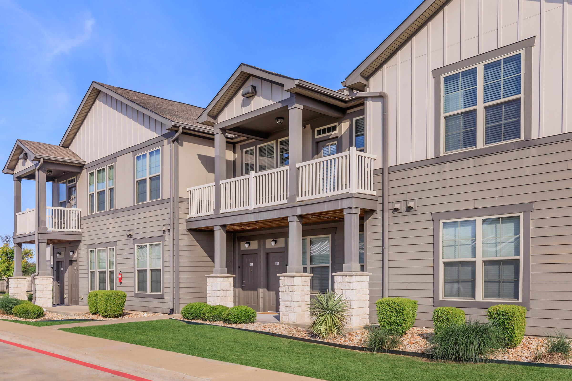 A modern two-story apartment building featuring a gray exterior with white accents, balconies, and landscaped shrubs. The structure includes multiple entrances and large windows, set against a clear blue sky.