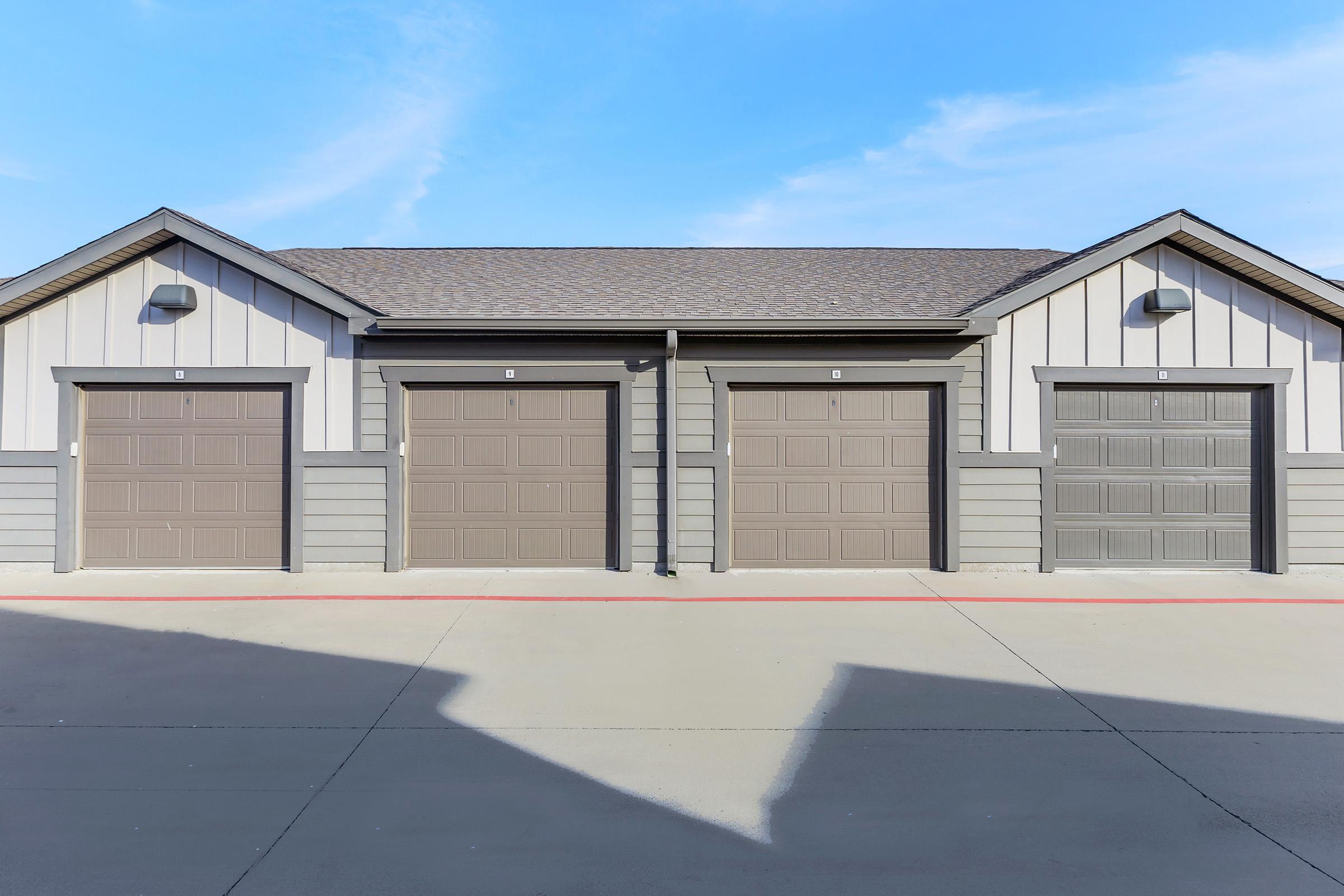 A view of a small storage facility featuring several beige garage doors. The building has a modern design with a gray and white color scheme, and a clear blue sky is visible above. The pavement in front is plain and unmarked, indicating a tidy and organized space.