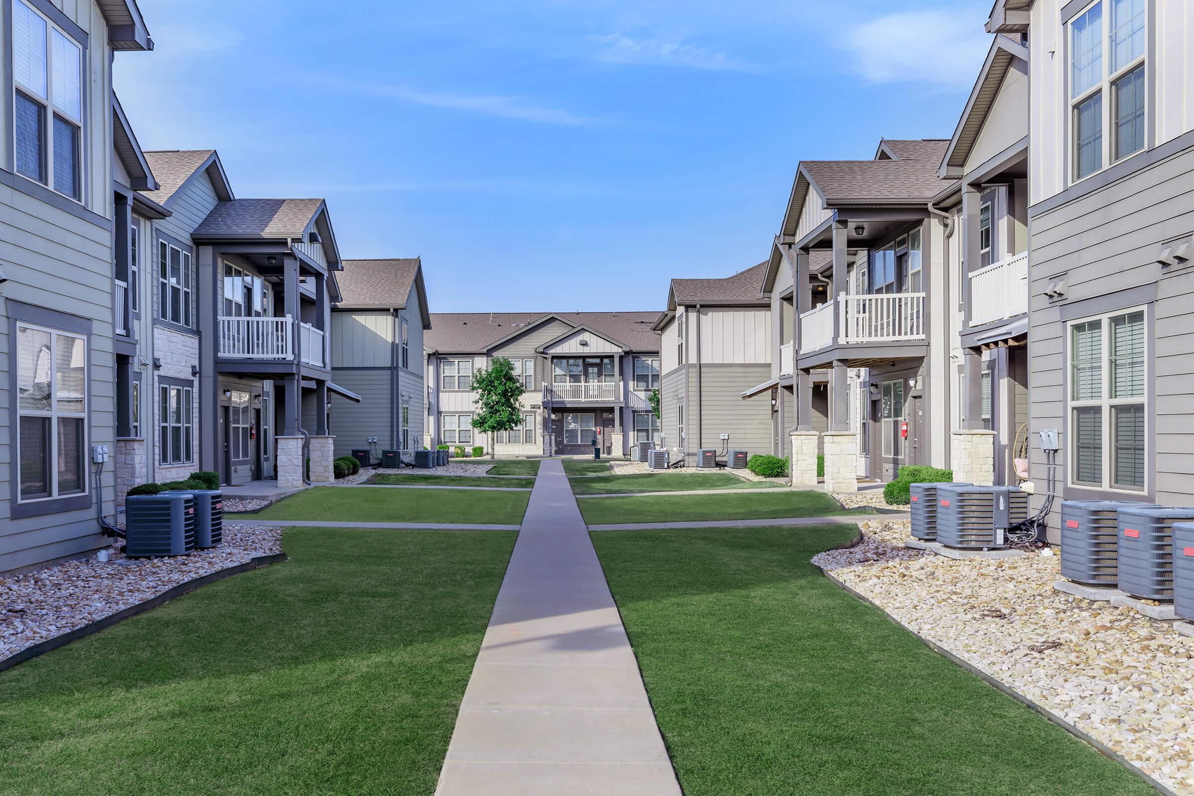 A spacious apartment complex featuring multiple buildings arranged around a landscaped courtyard. The pathway leads through neatly maintained grass and gravel areas, with air conditioning units visible near the buildings under a clear blue sky.