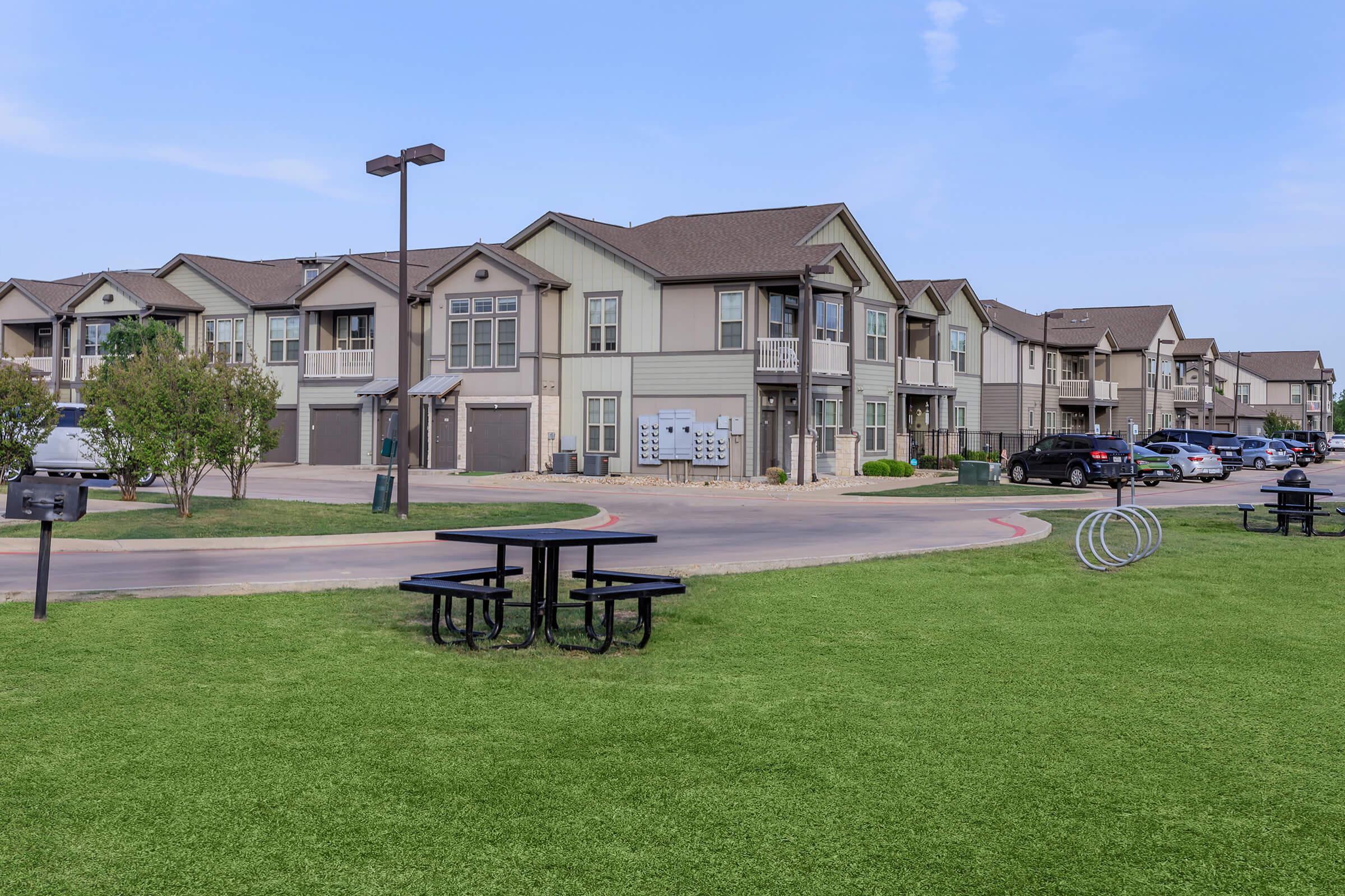 A residential apartment complex featuring multiple buildings with modern architecture. There are picnic tables on a green lawn, BBQ grills nearby, and a bike rack. The area is well-maintained, with asphalt pathways and parking spaces for vehicles. The sky is clear with a few clouds.