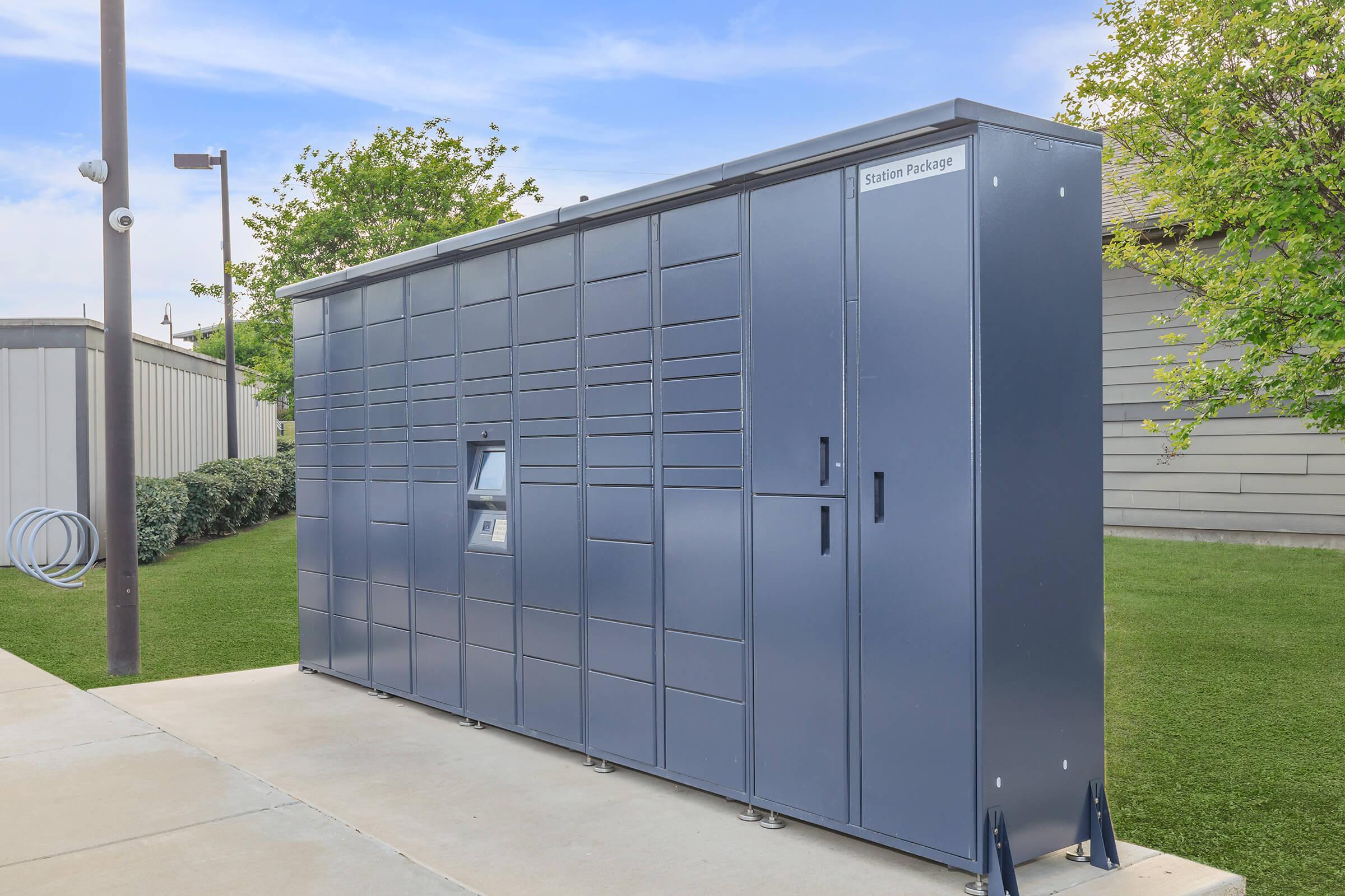 A large, modern package locker system situated outdoors on a paved surface. The locker is gray and features multiple compartments for package delivery, with a small access panel for retrieving items. Surrounding greenery and a light pole are visible in the background.