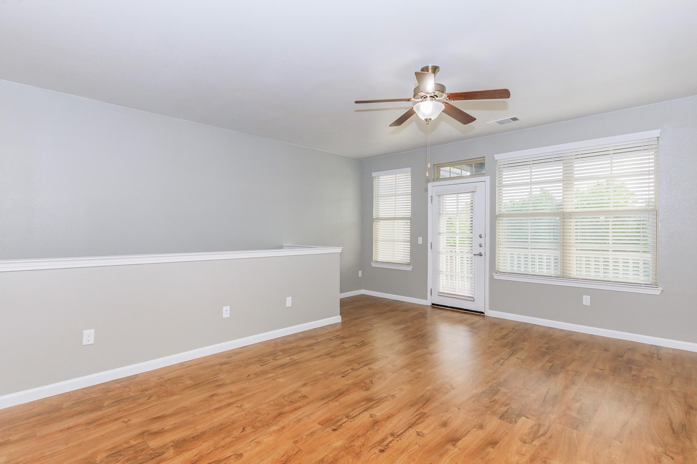 A bright living room featuring light gray walls, a ceiling fan, and large windows with white blinds. The floor is made of warm wood laminate, and a doorway leads to a small balcony. The space is open and inviting, perfect for relaxation and natural light.