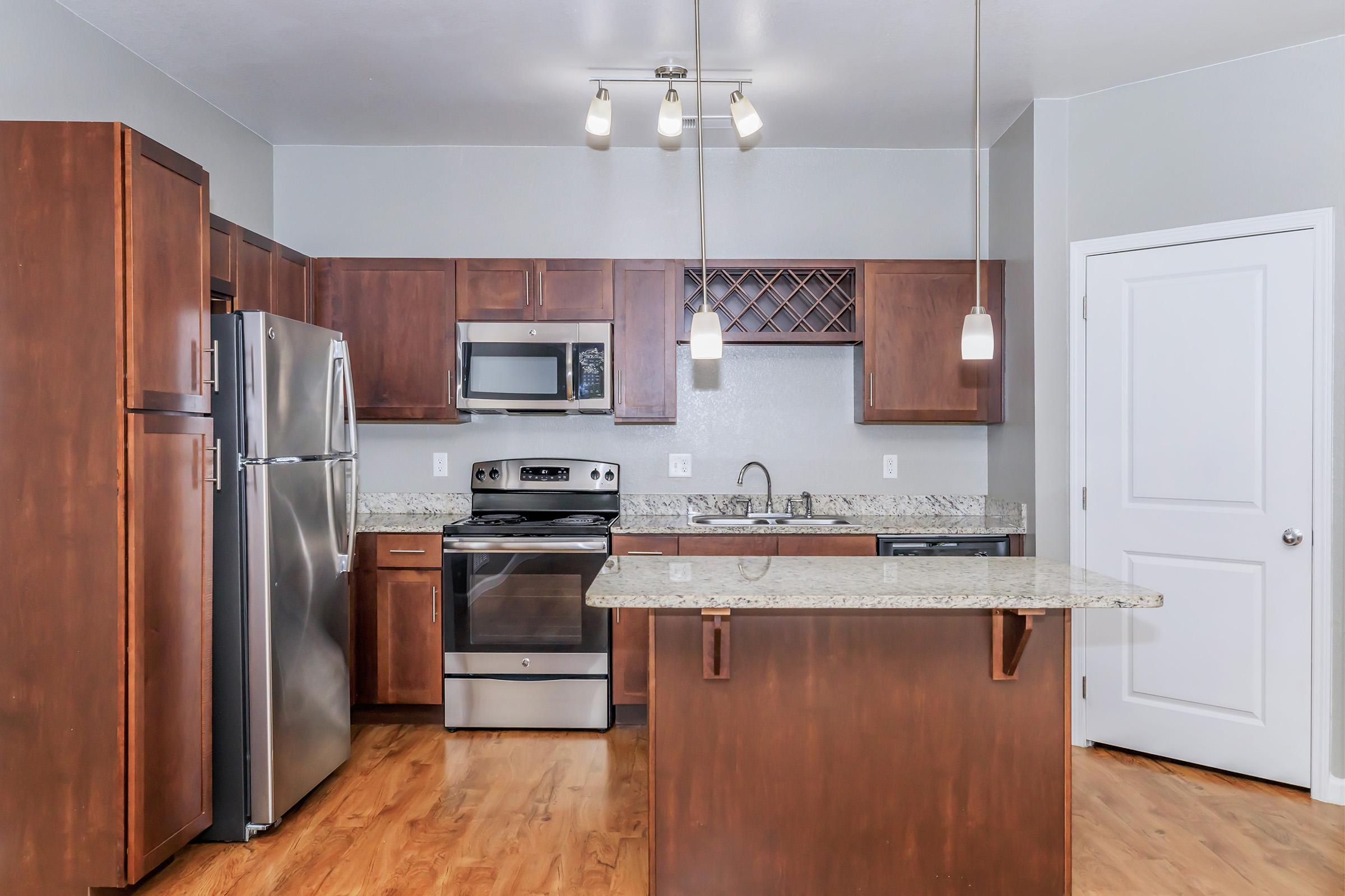 Modern kitchen featuring dark wood cabinets, stainless steel appliances, and a granite countertop island. The space includes pendant lighting and a wine rack, with beige walls and a wooden floor, creating a warm and inviting atmosphere.