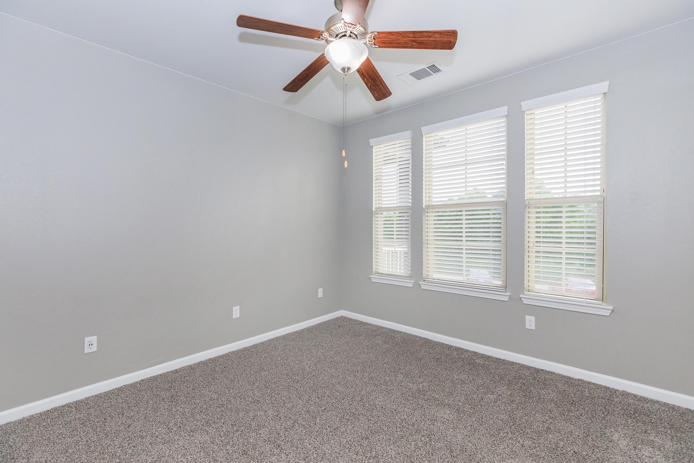 A well-lit room with gray walls and a ceiling fan. The floor is covered with plush carpet, and there are three windows with white blinds that allow natural light to enter.